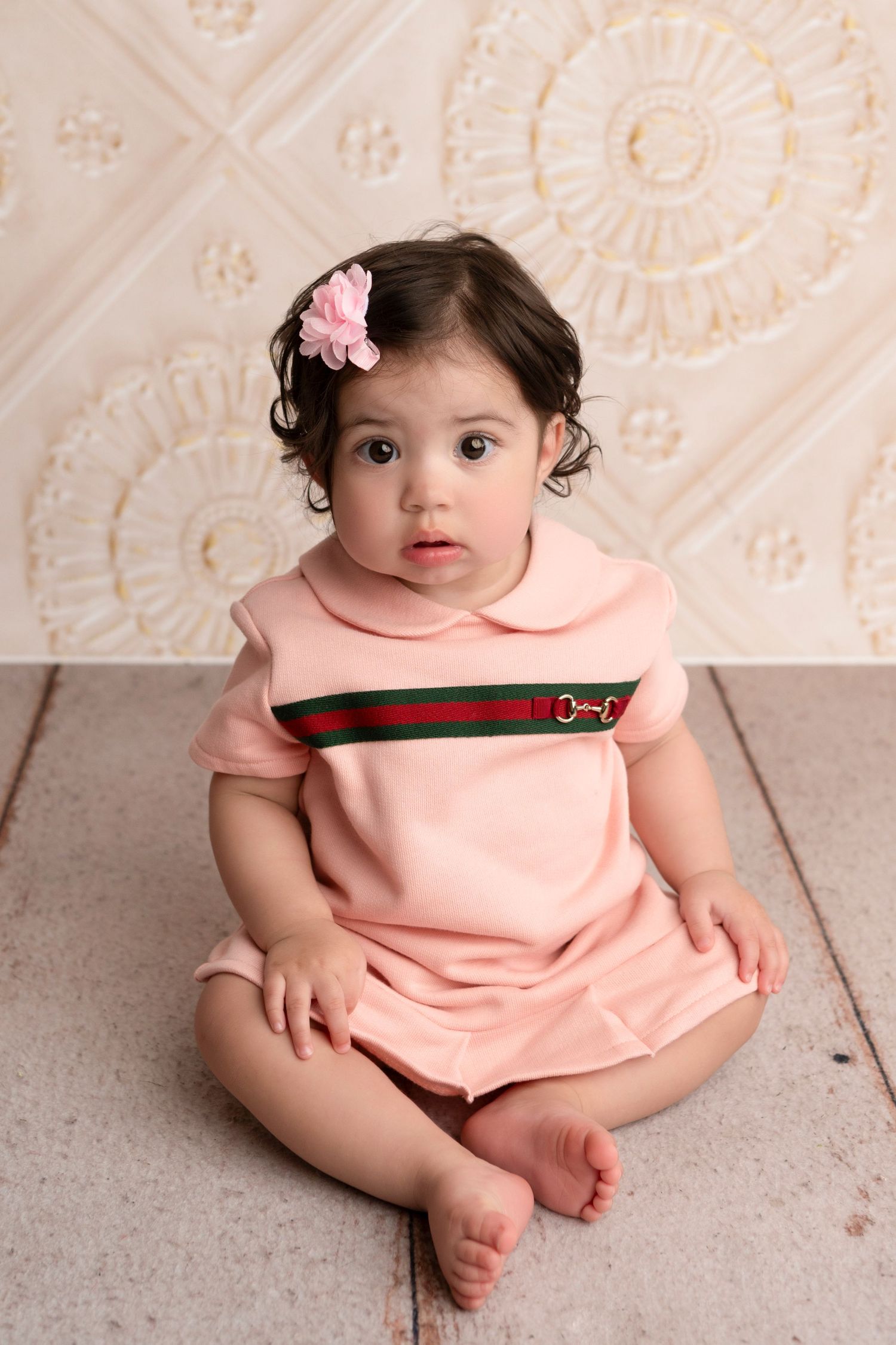 A toddler in a pink collared dress with a flower hair clip sits against a textured cream background.