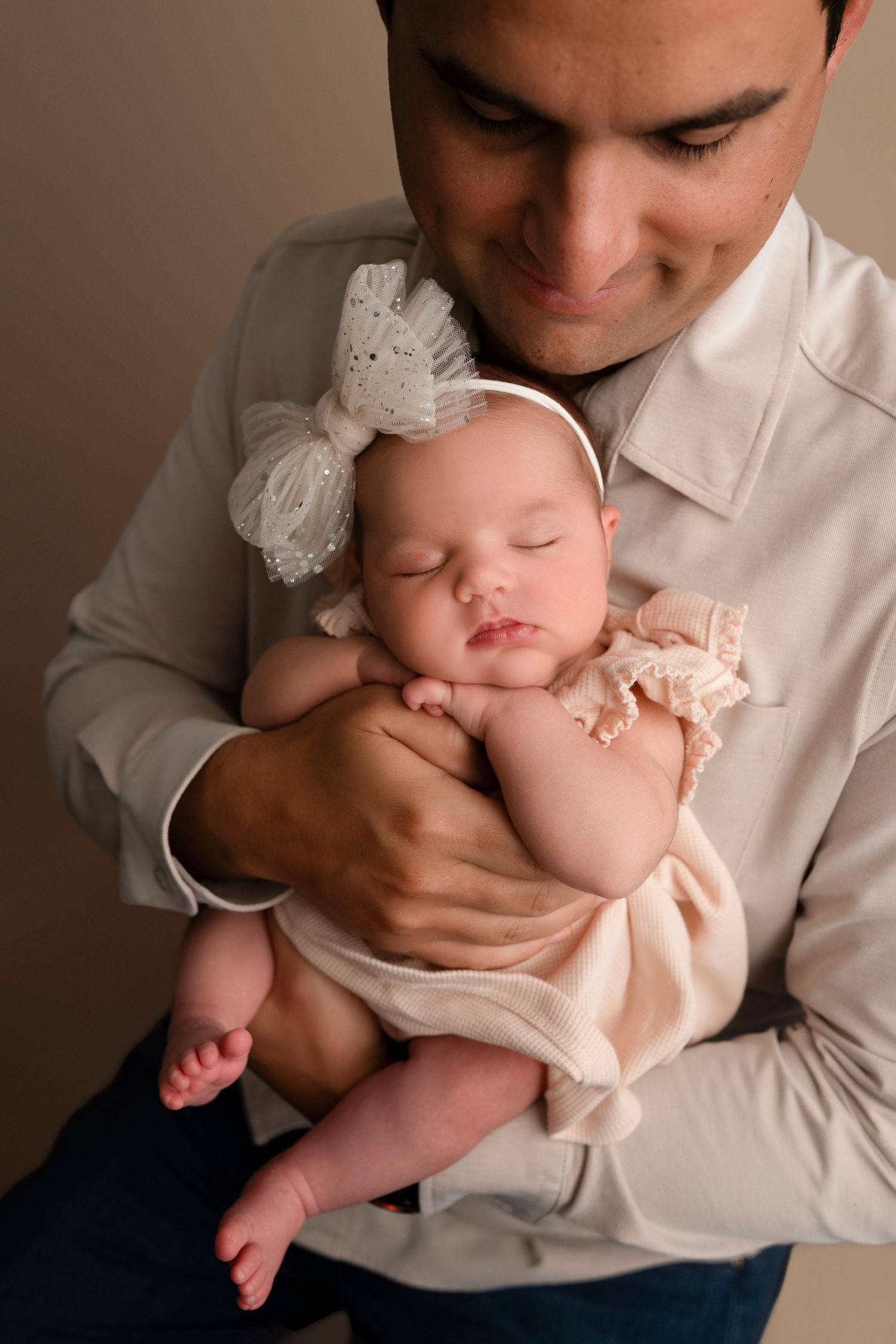 A sleeping newborn baby wearing a white bow headband rests peacefully in caring arms against a neutral background.