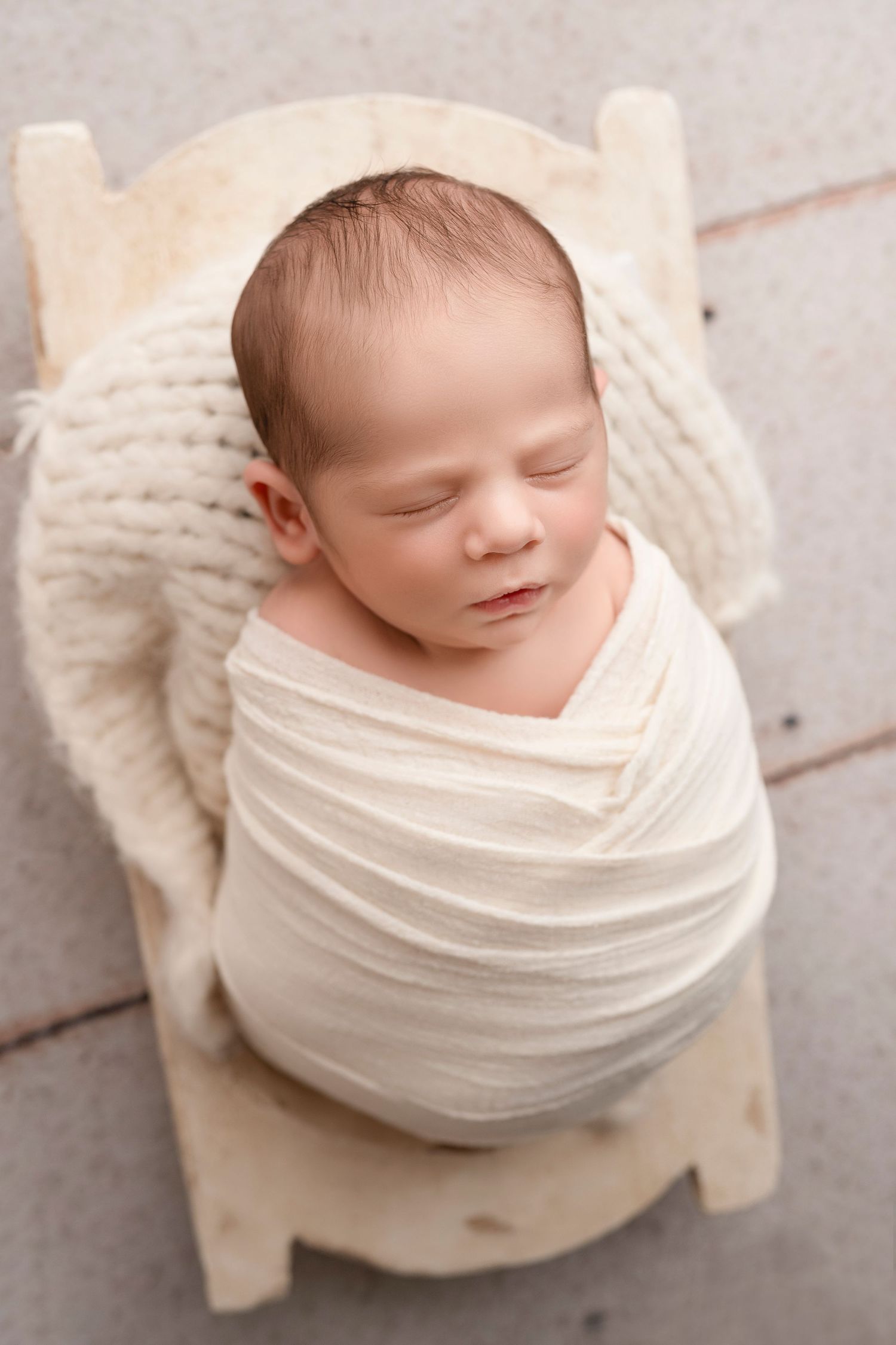 Sleeping newborn wrapped in cream fabric lying on small wooden bed with knitted blanket.