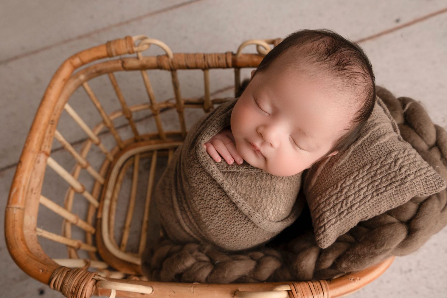 Peaceful sleeping newborn wrapped in brown swaddle blanket rests in curved wooden basket.