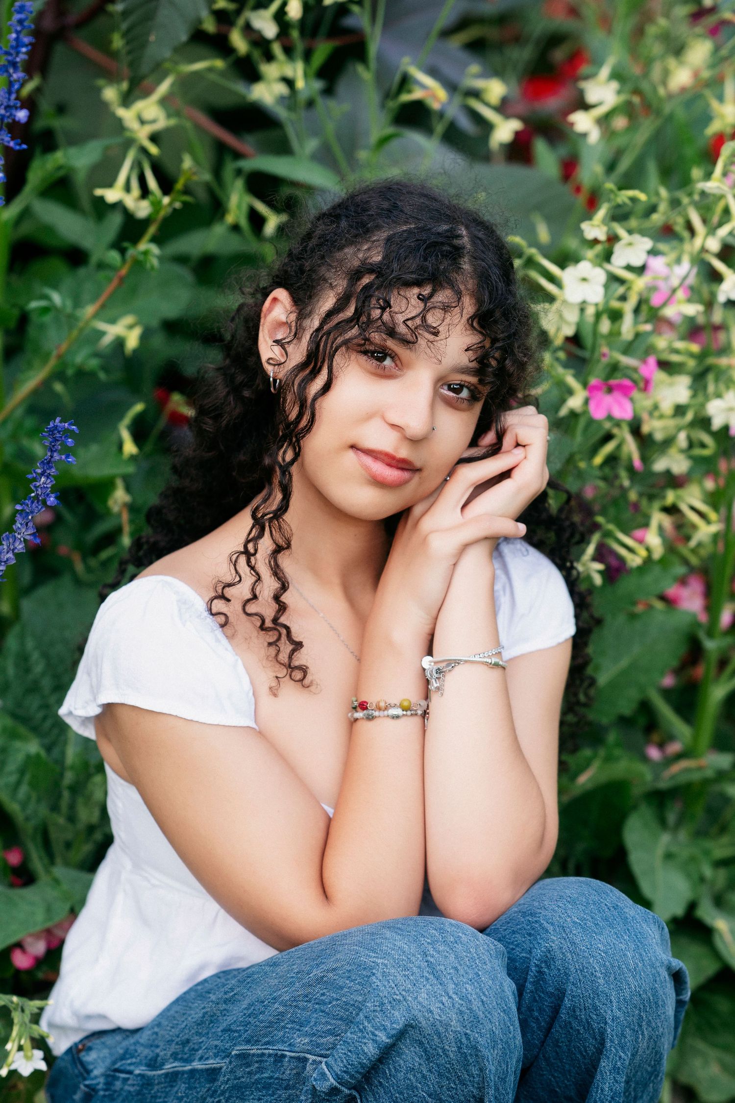 A person in a white top and jeans poses outdoors against a lush green garden backdrop with colorful flowers.