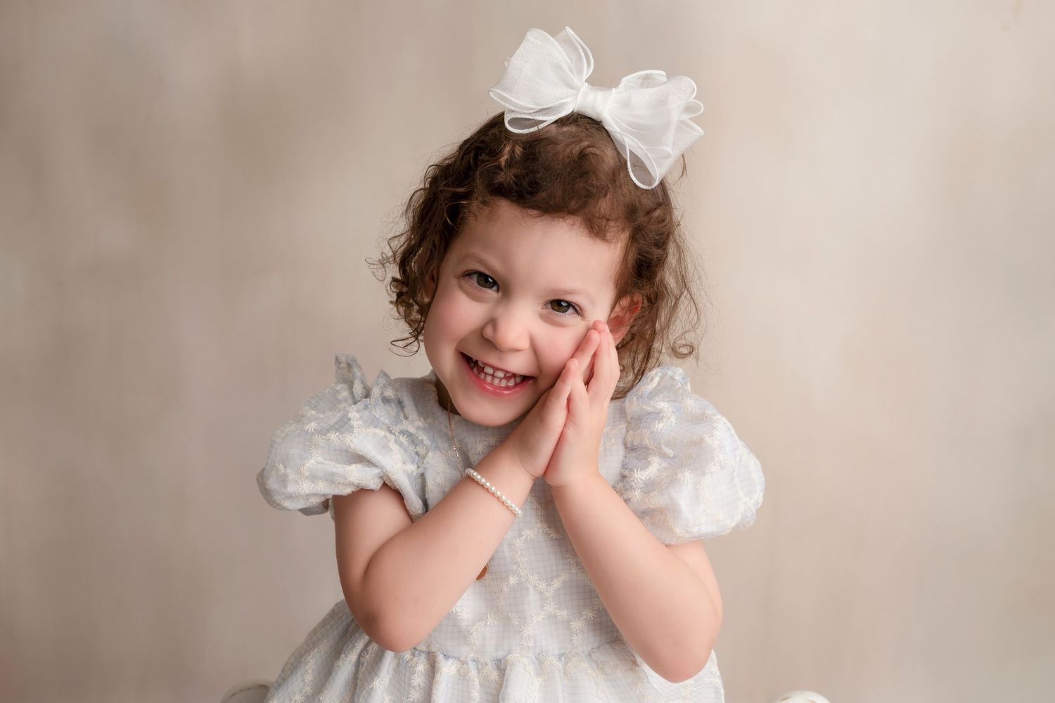 A child in a white vintage dress poses playfully against a neutral backdrop in a portrait studio setting.