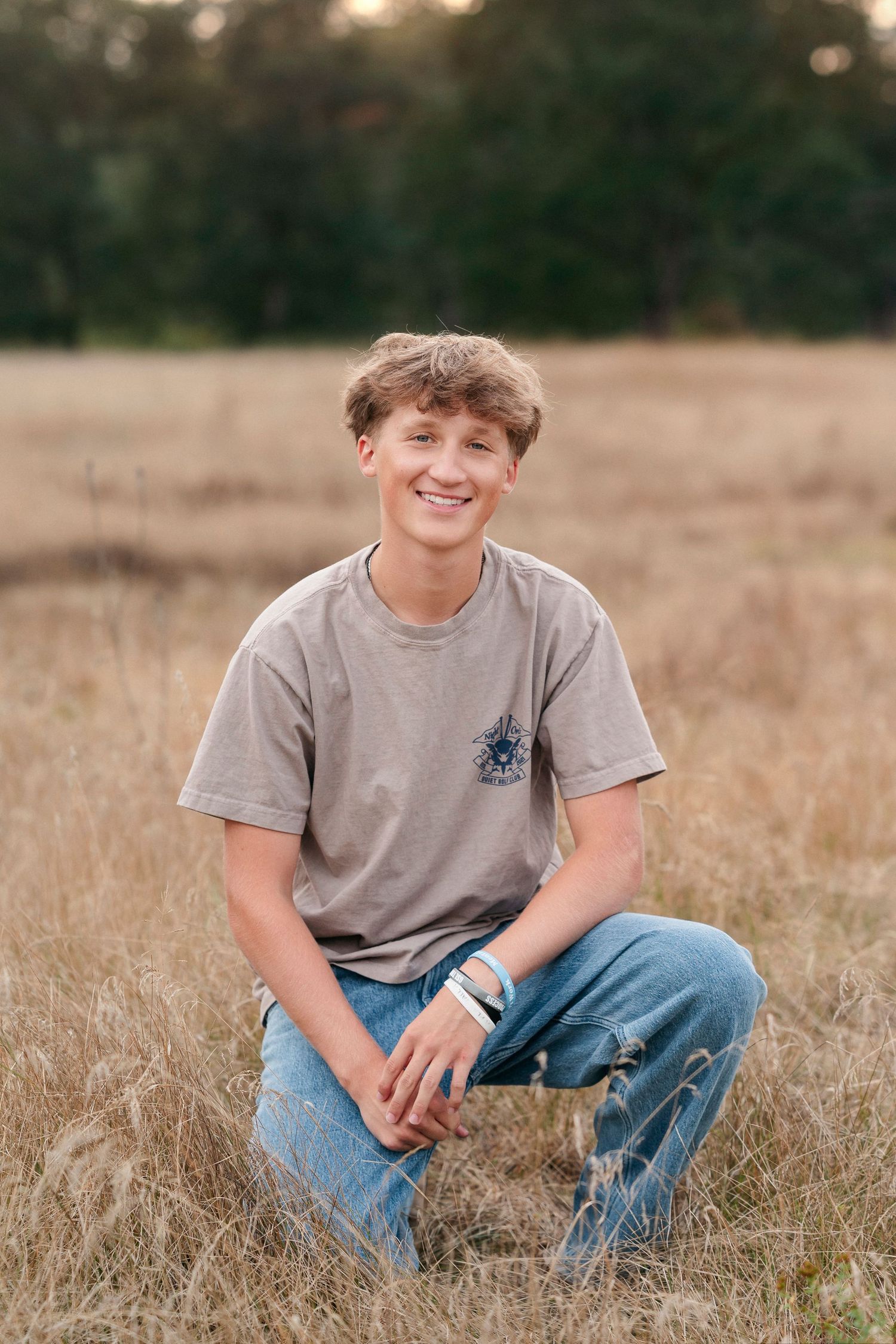 Person in gray t-shirt and jeans kneeling in a golden field at sunset with trees in background.