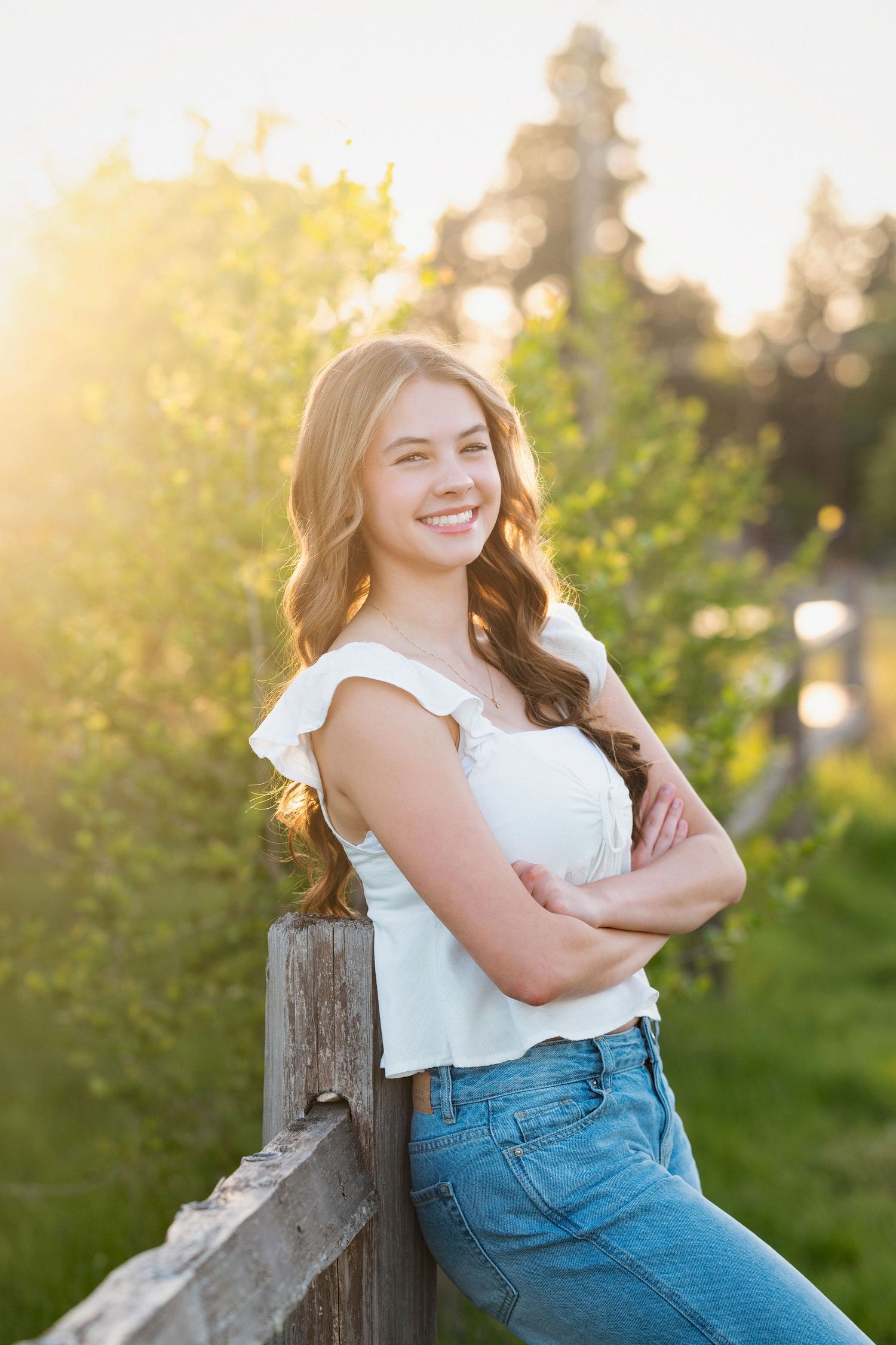 A person in a white top and denim sits casually on a wooden fence during golden hour with greenery in the background.