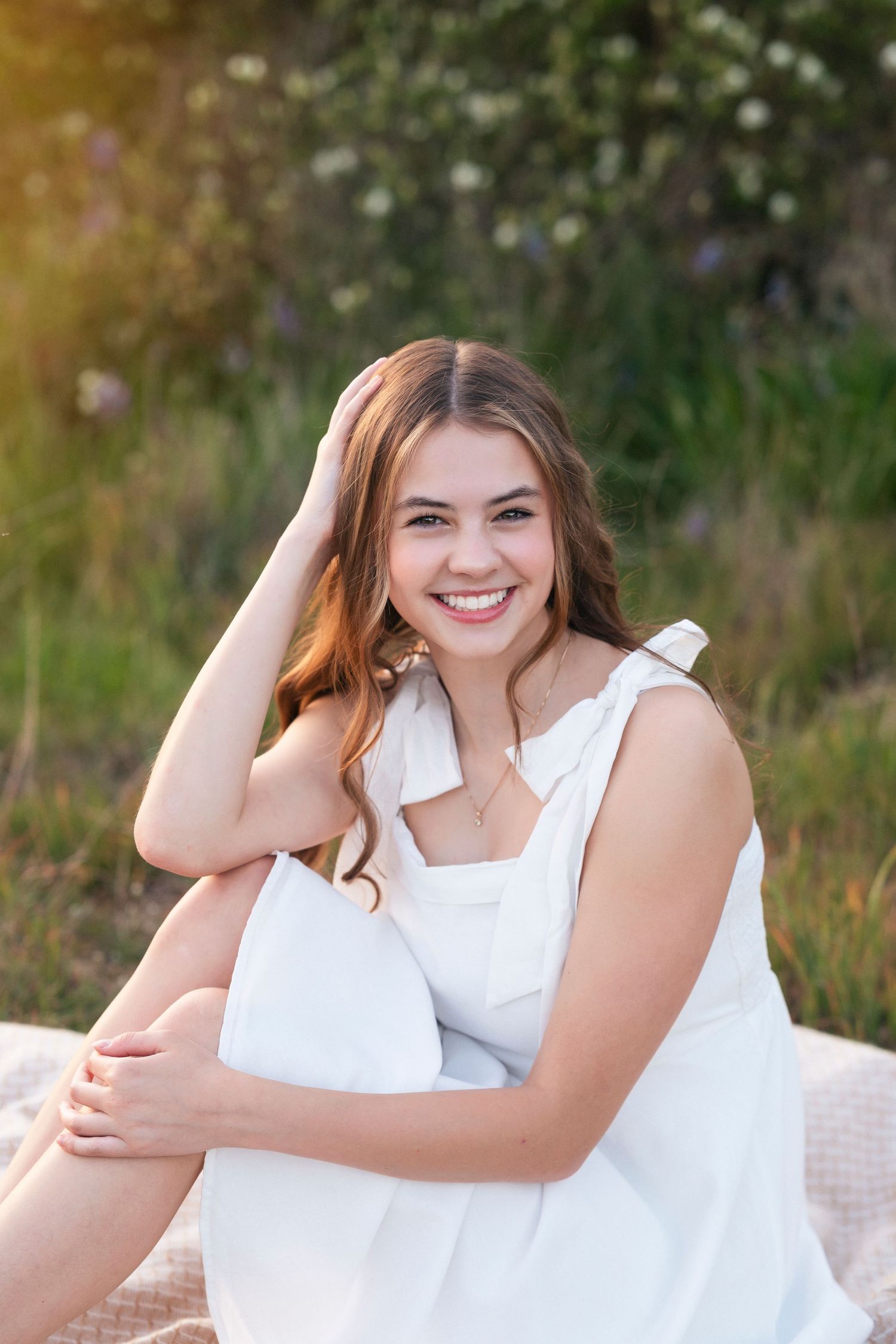 A smiling person in a white summer dress sits outdoors on a blanket with sunlit greenery in the background.
