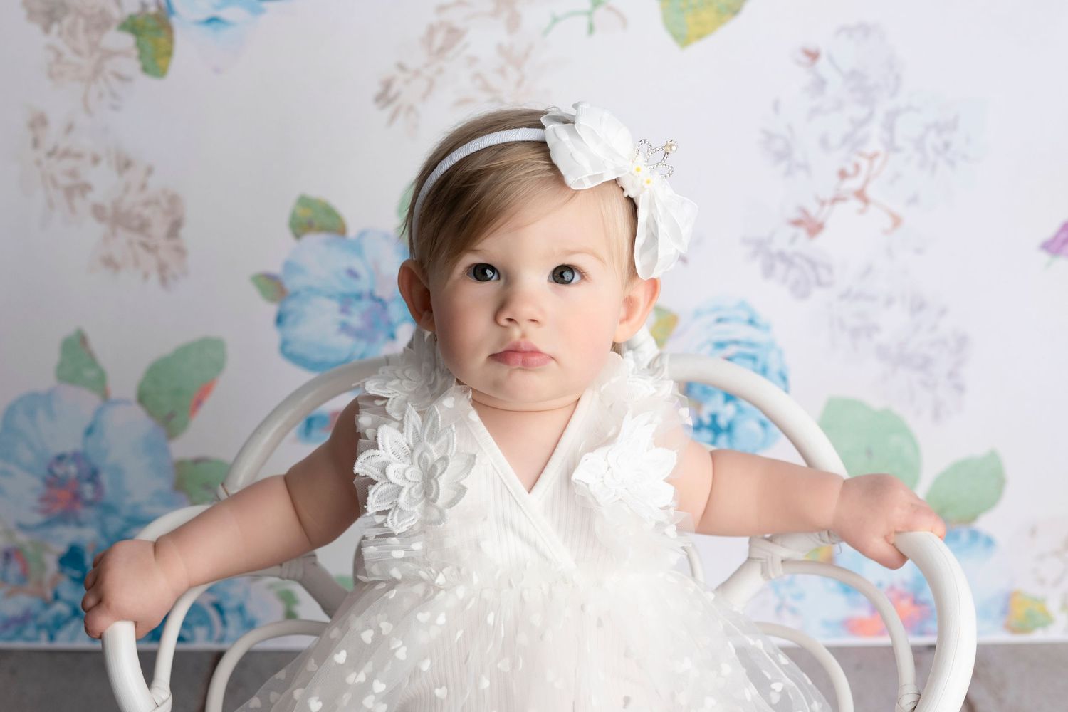 A baby girl in a white ruffled dress and bow headband sits in a white chair against a floral backdrop.
