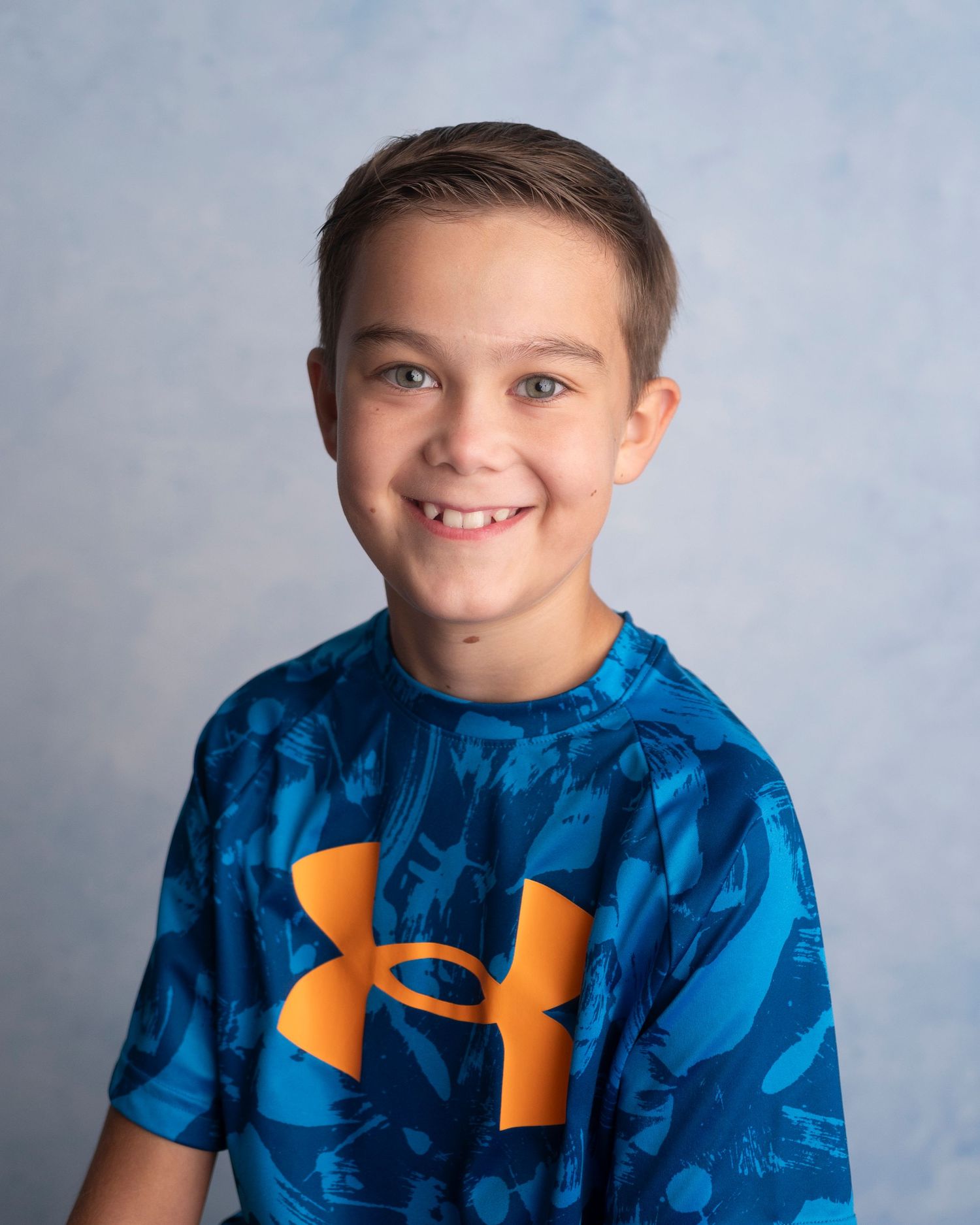 Smiling young boy wearing a blue Under Armour t-shirt, posing for a school portrait photo against a gray background.