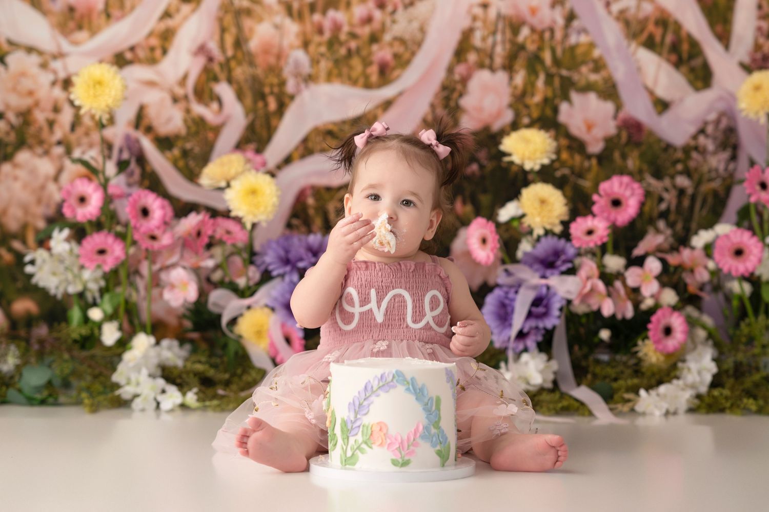 Baby girl in pink tutu eating cake at her first birthday cake smash with floral backdrop.