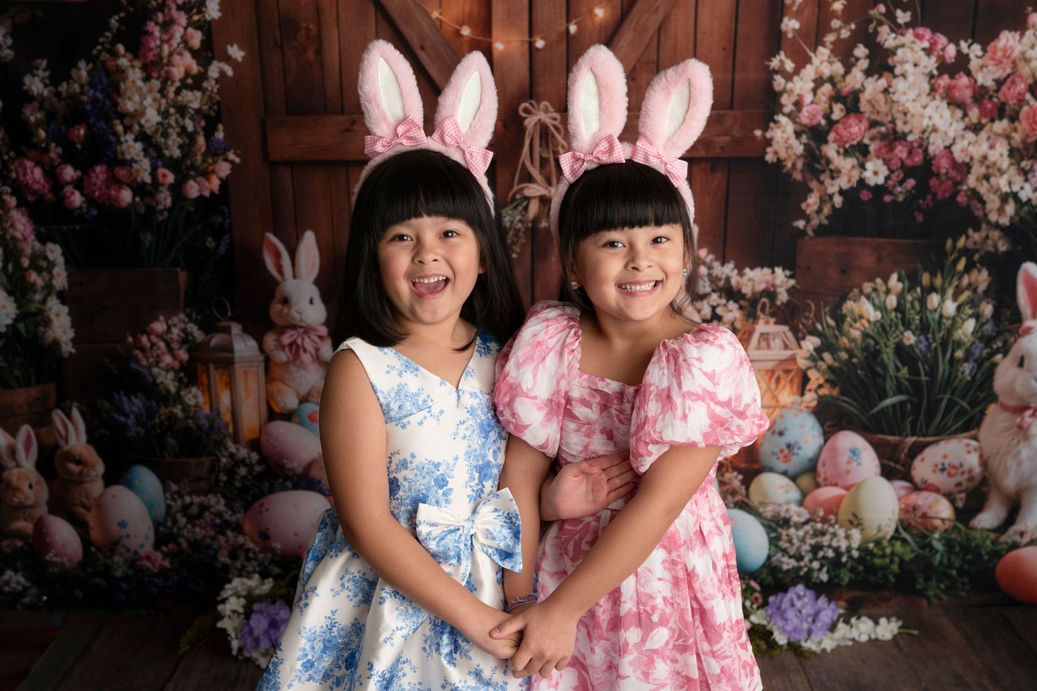 Two young girls in floral dresses and bunny ears pose with Easter eggs and bunnies in a festive spring photo.