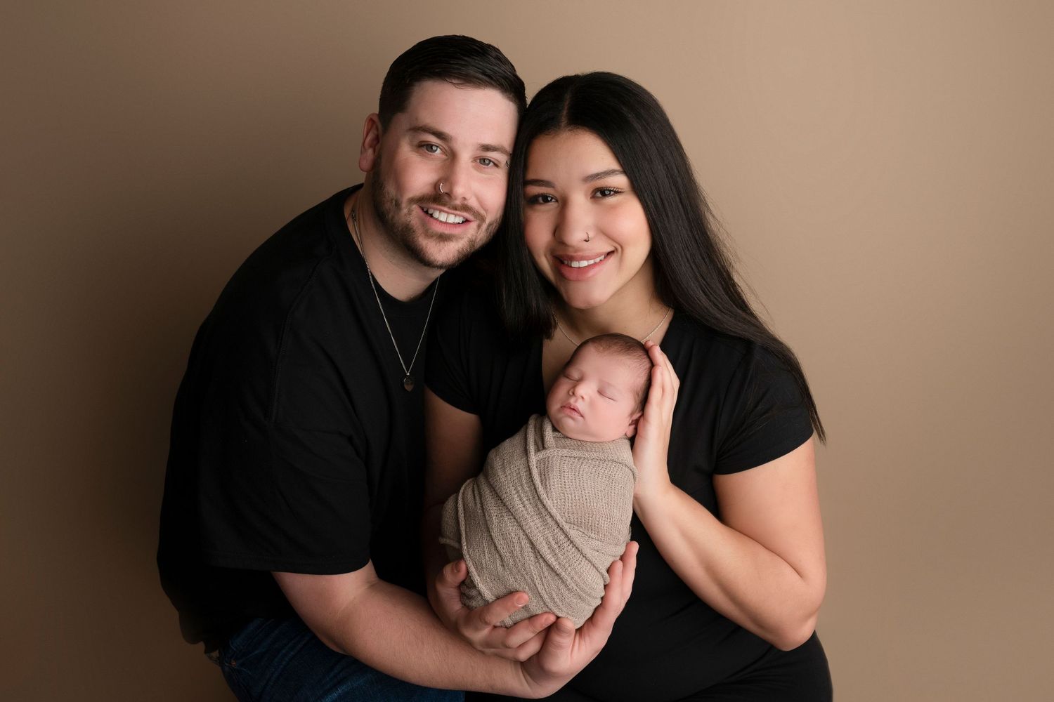 A smiling couple in black shirts poses with their swaddled newborn baby against a warm beige studio backdrop.