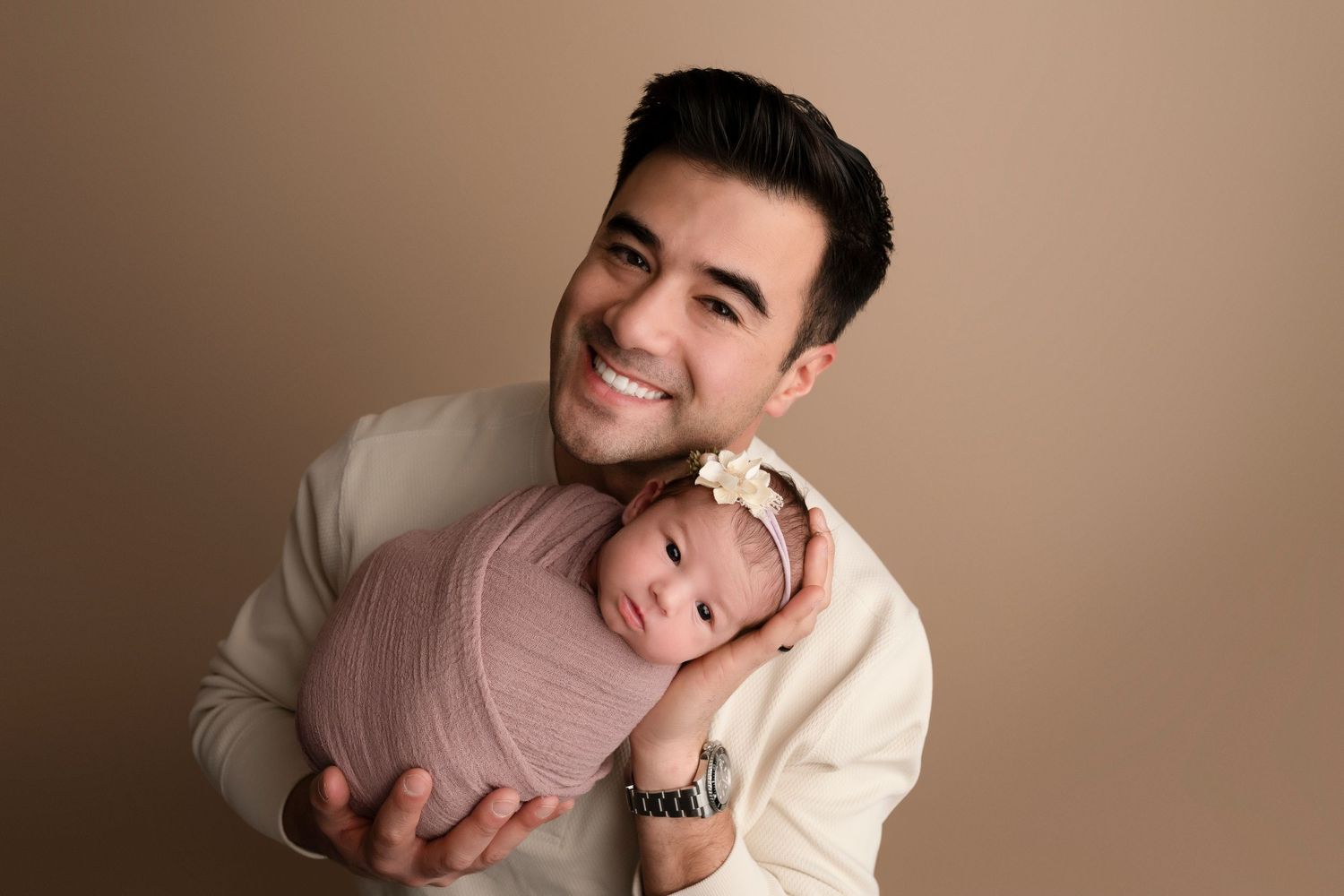Smiling father holds swaddled newborn baby in pink wrap with floral headband against beige studio backdrop.