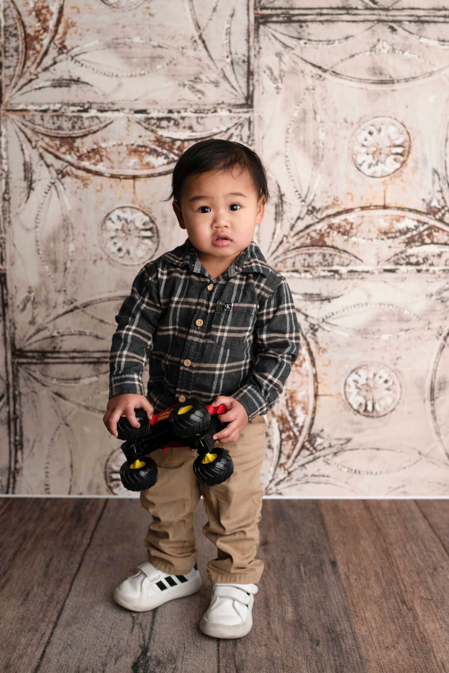 Toddler boy in plaid shirt and khaki pants holding a toy monster truck, standing in front of decorative backdrop.