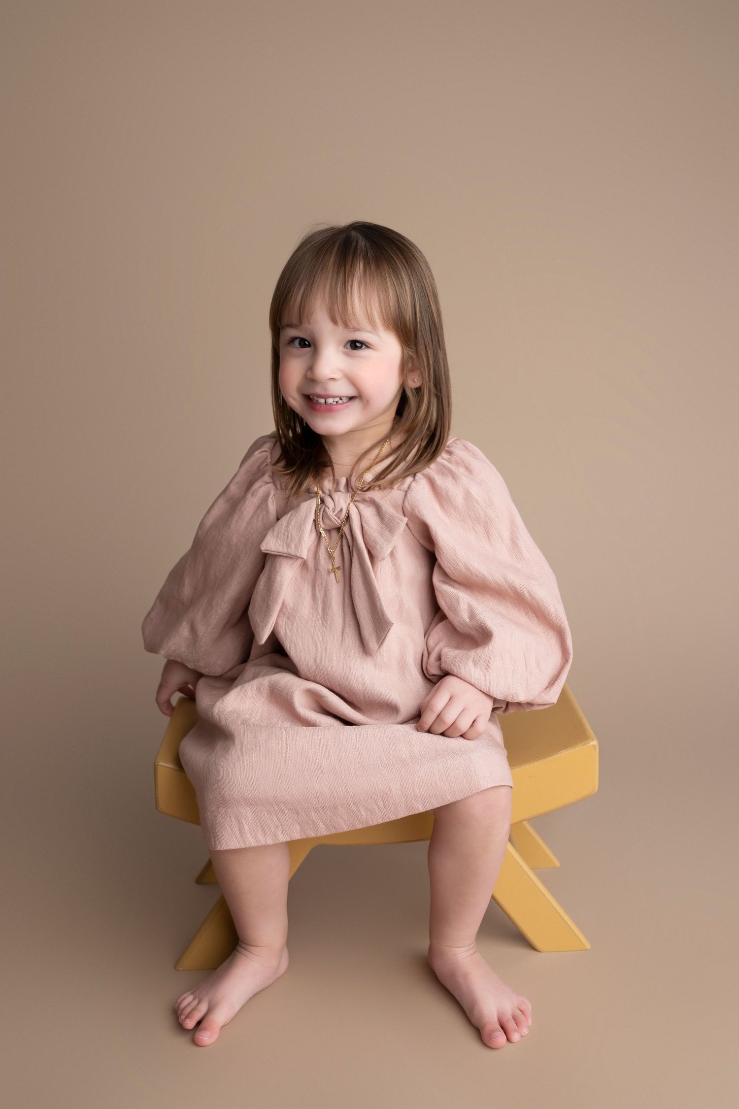 A smiling young girl in a pink linen dress sits barefoot on a yellow stool against a warm beige background.