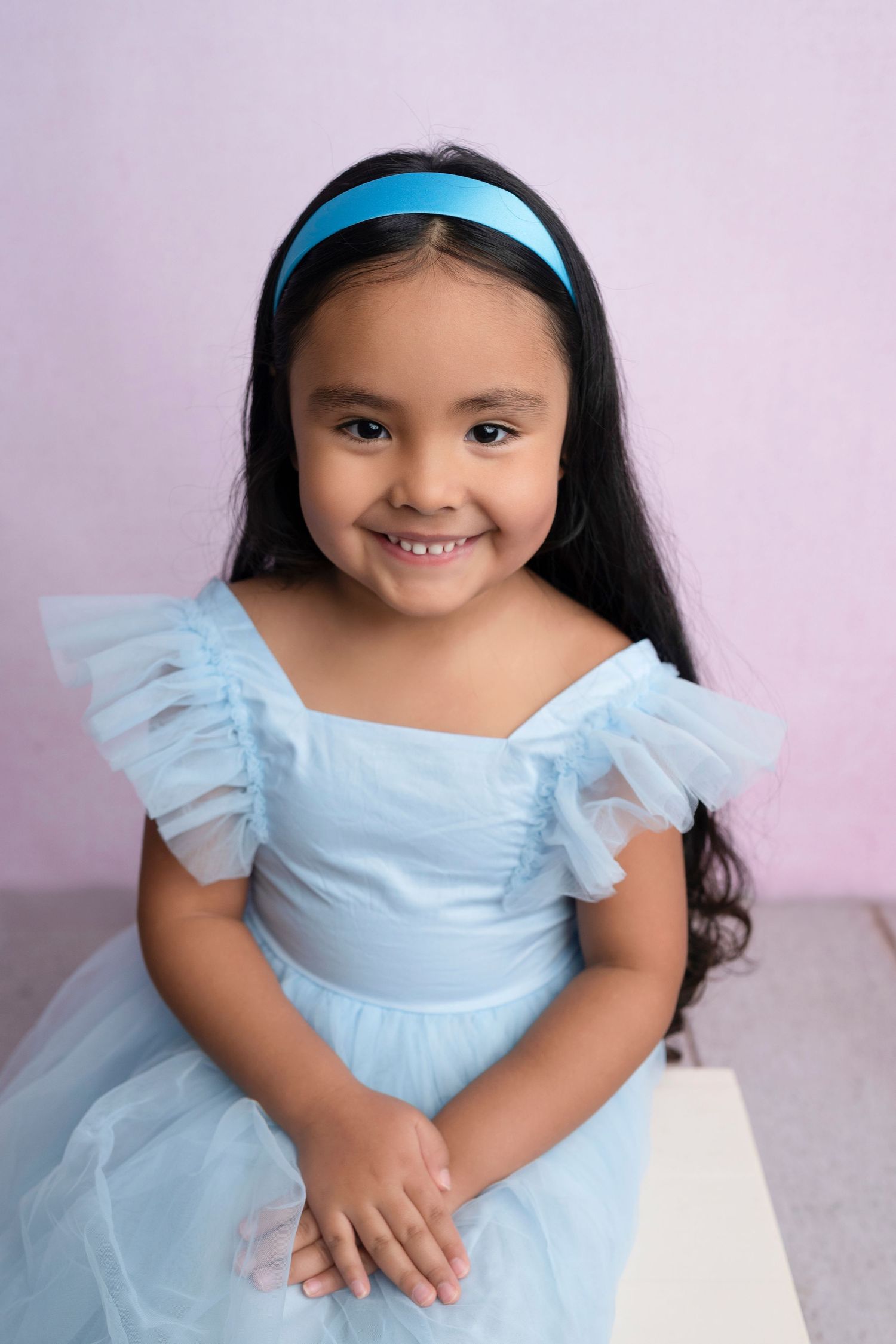 A smiling young girl in a light blue tulle dress and matching headband poses against a pink background.