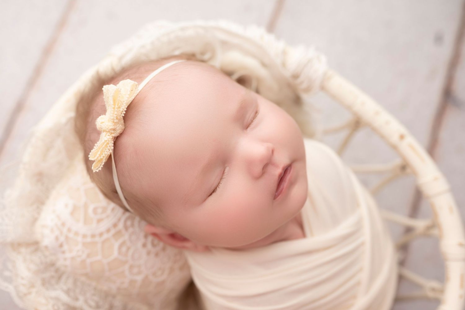 Sleeping newborn baby girl wrapped in cream lace swaddle with bow headband in white wicker basket.