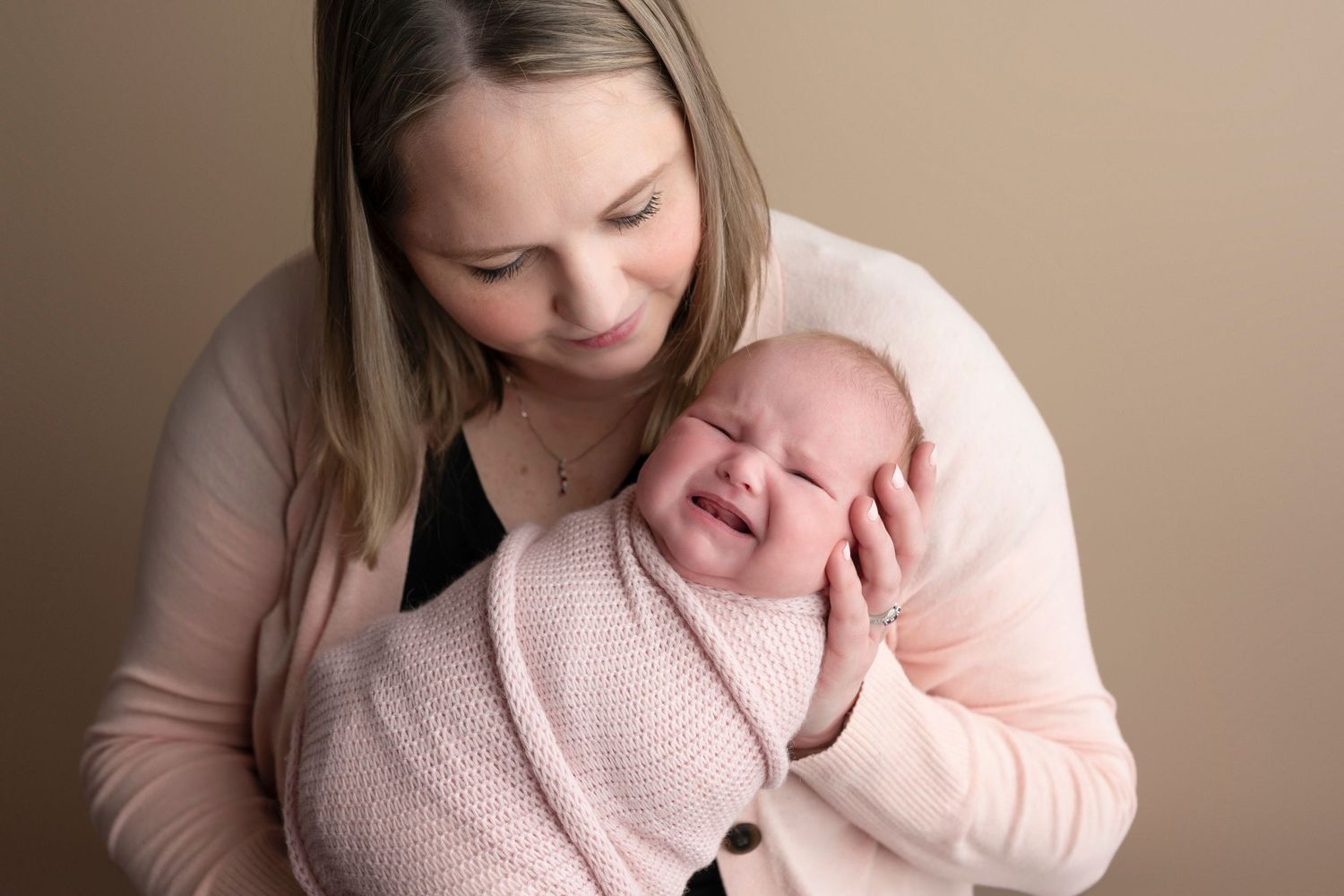 A mother tenderly holds her smiling newborn wrapped in a pink knit blanket against a warm beige background.