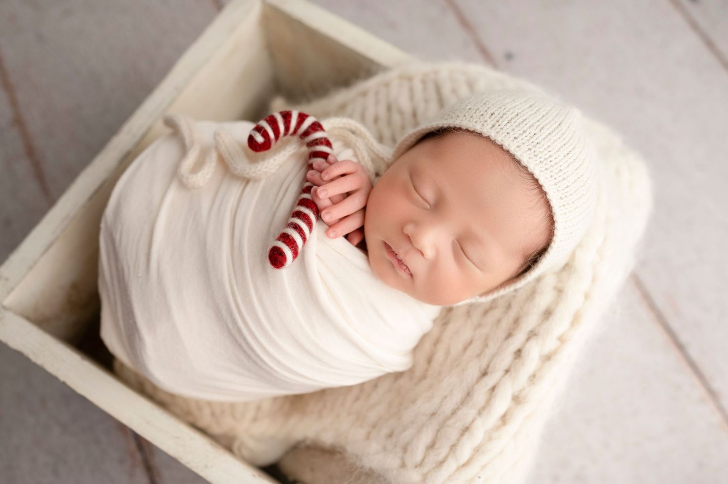 Sleeping newborn baby wrapped in white swaddle wearing knit bonnet, holding candy cane striped toy in wooden box.