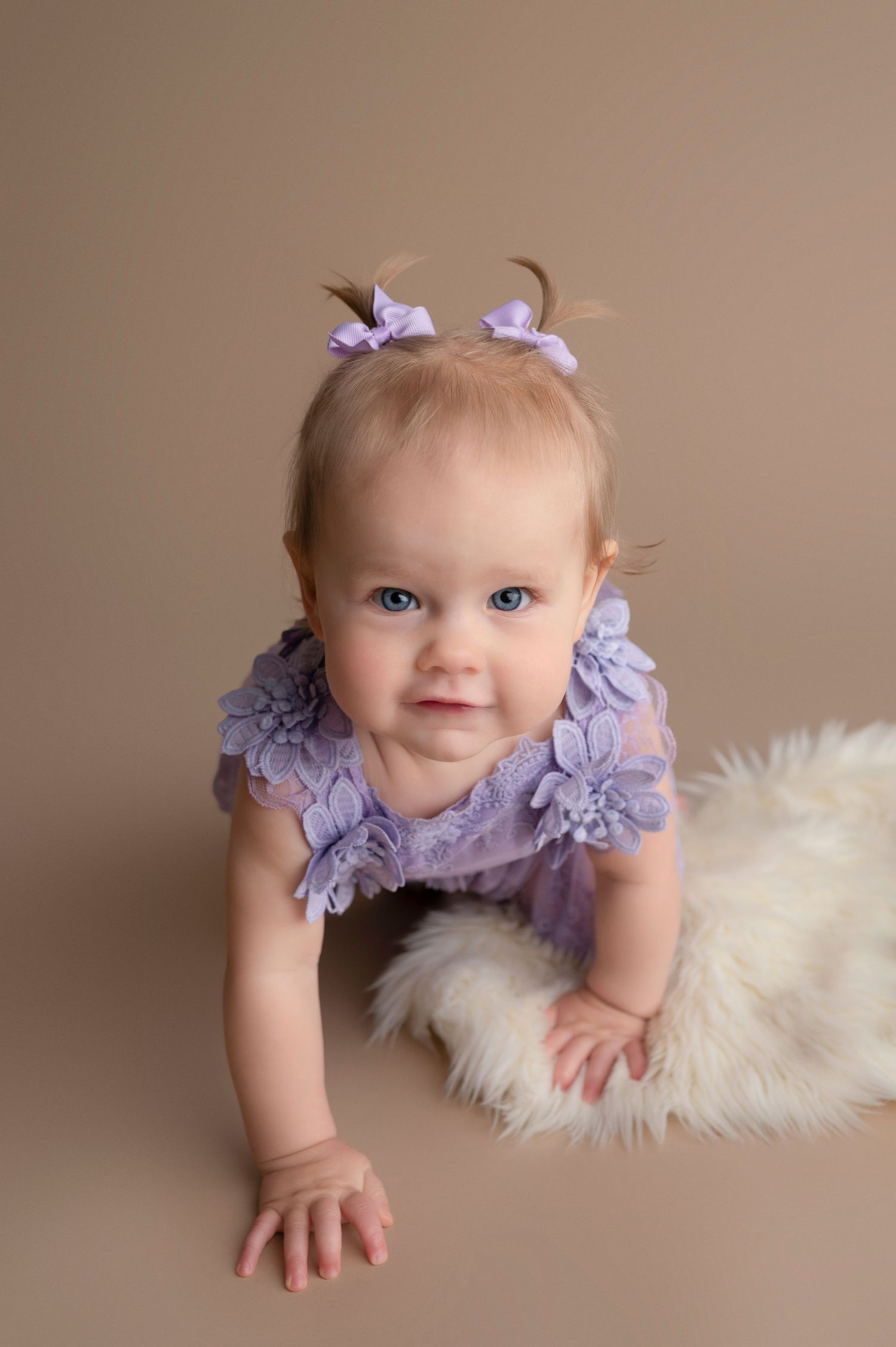 Adorable baby girl in purple floral romper with pigtail bows crawling on a fluffy white rug.