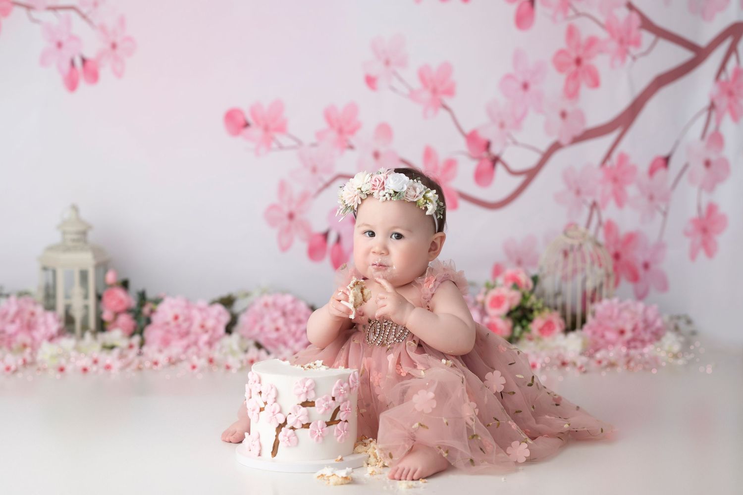 Baby girl in pink tutu dress sits before floral cake amid cherry blossom backdrop for milestone photo.