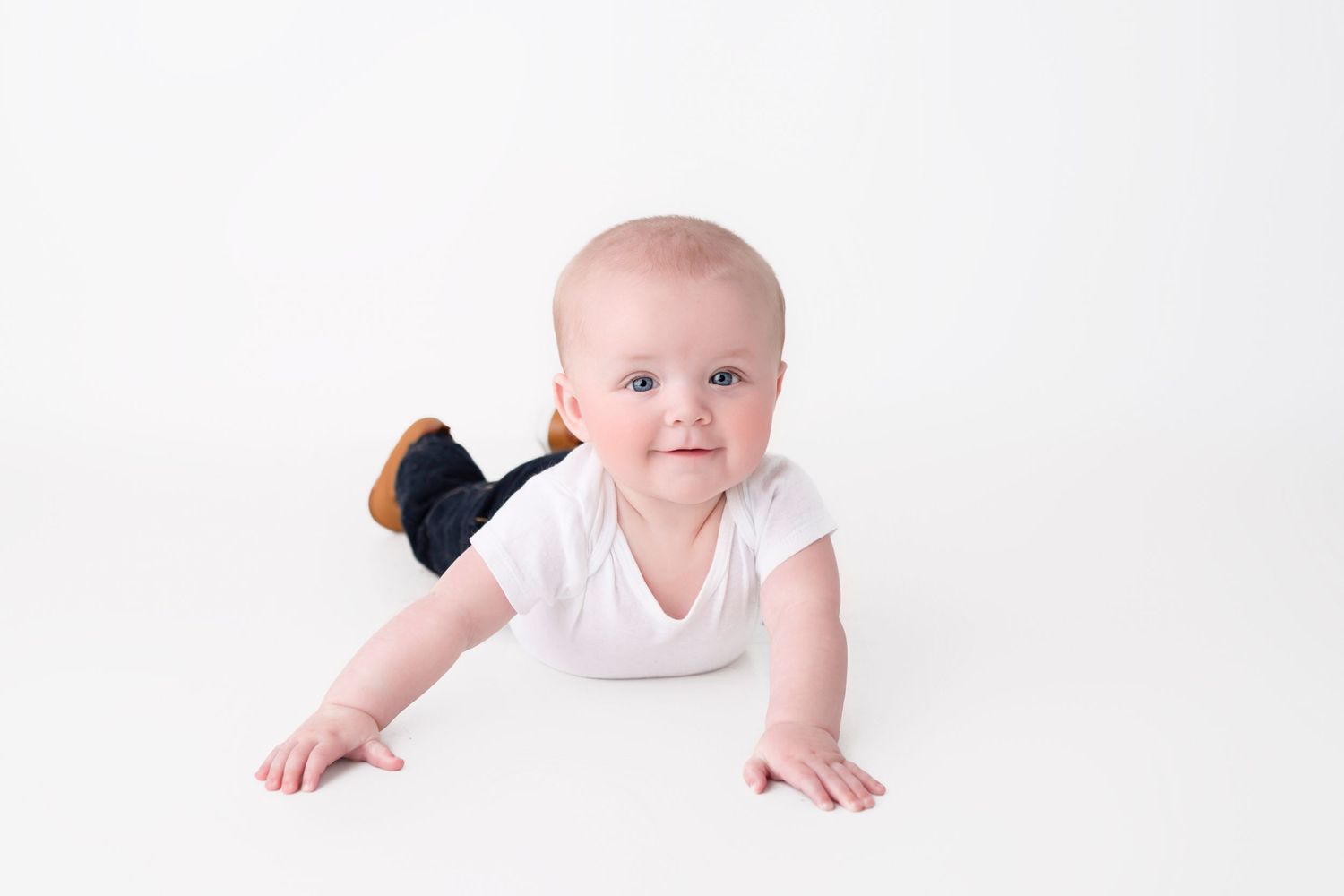 Adorable baby in white onesie and dark overalls crawling on white background, looking up with bright blue eyes.