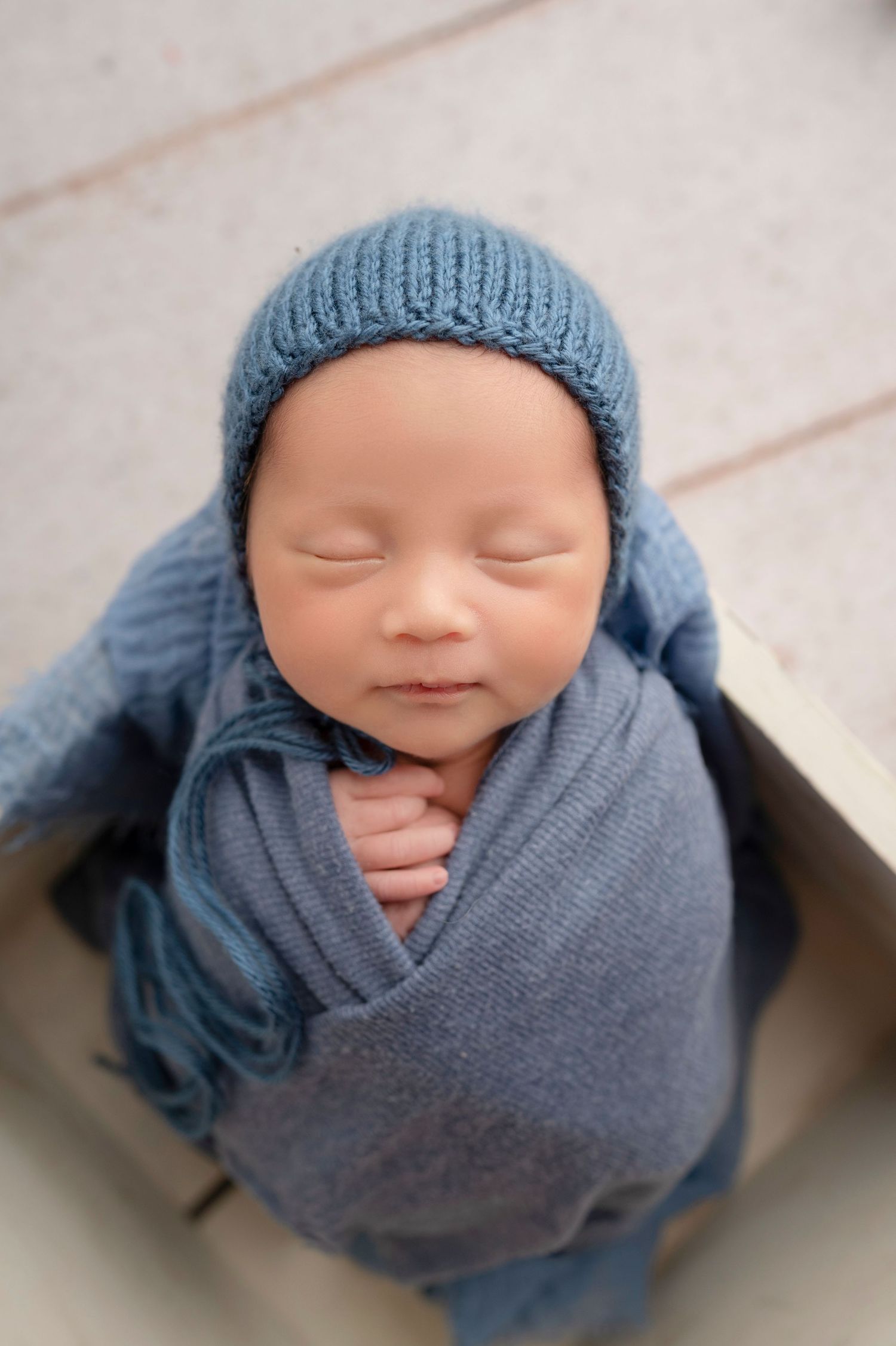 Sleeping newborn baby wrapped in a soft blue knit blanket and matching bonnet hat, nestled in a wooden box.