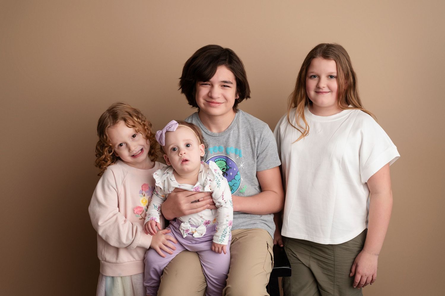 Four children pose together smiling in a studio portrait against a warm beige background.