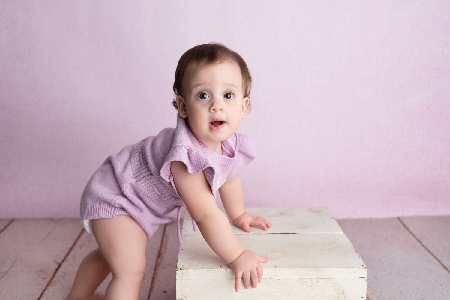 Baby in lavender outfit crawls and plays on white wooden floor against pink wall backdrop.