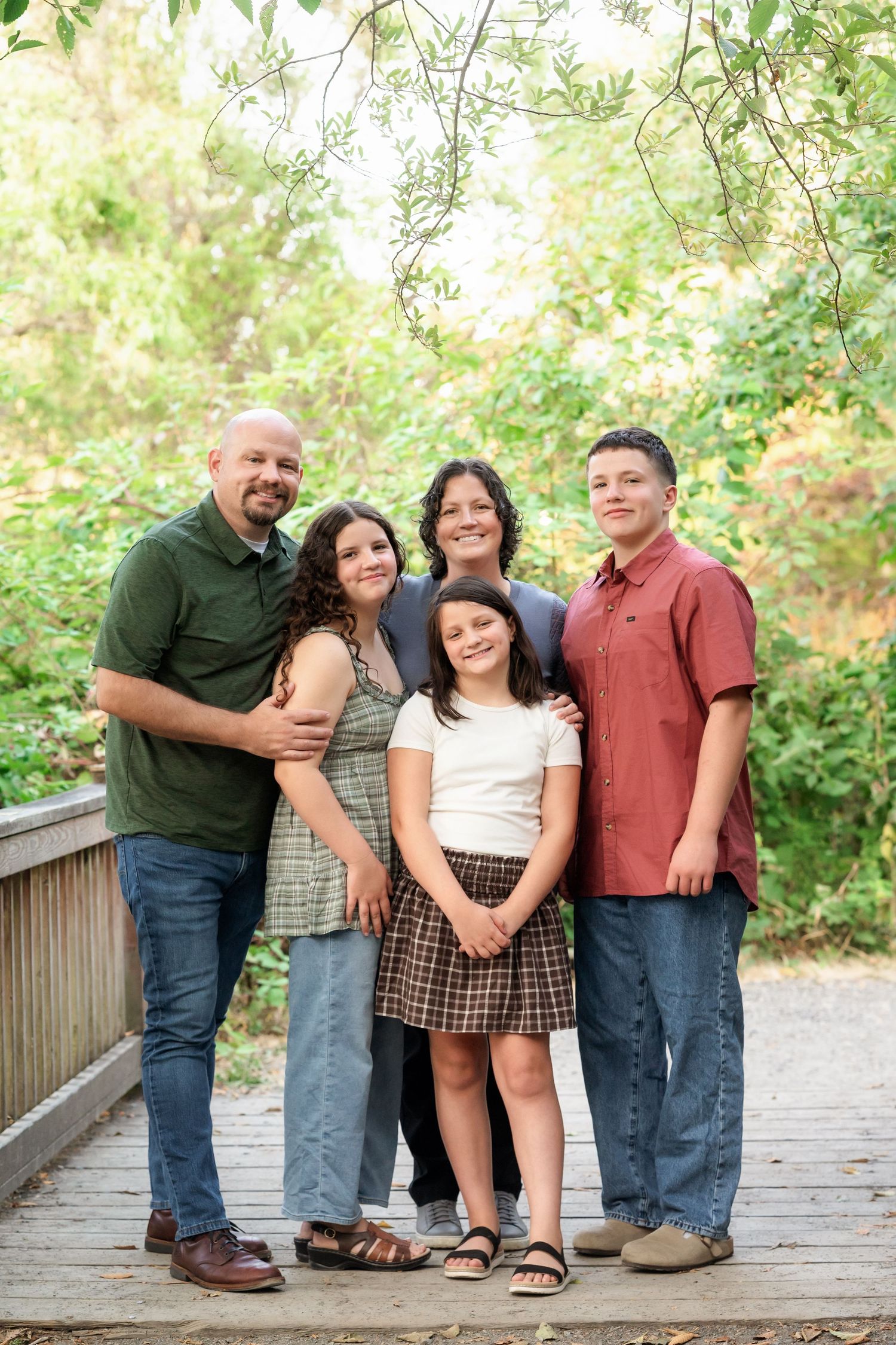 Family of five posing together on a wooden bridge surrounded by lush green trees outdoors.