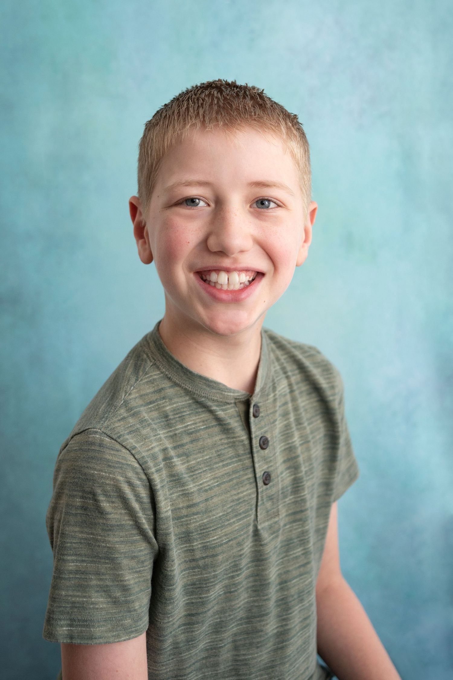 Smiling preteen boy with short blonde hair wearing a gray henley shirt posed against a blue background.