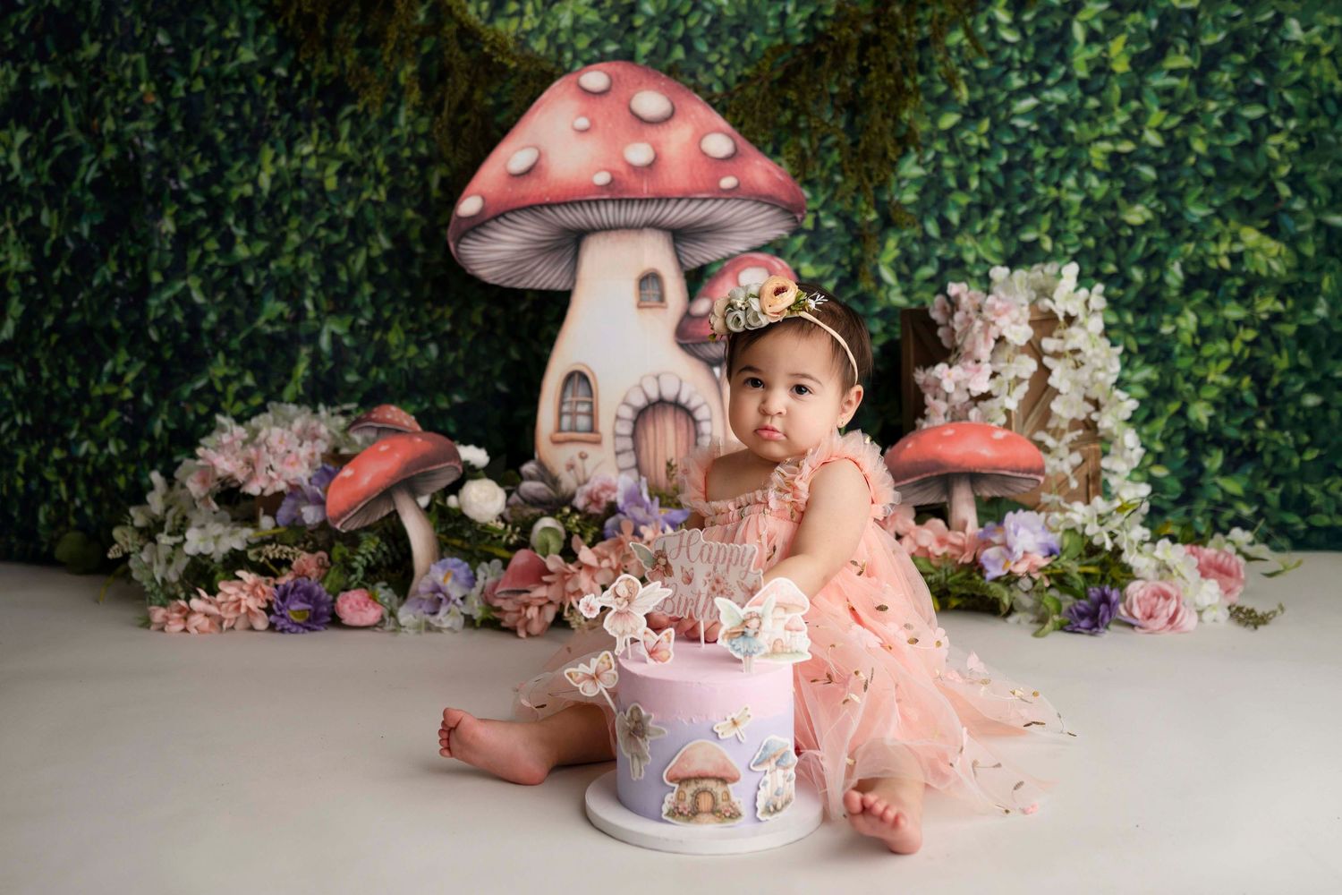 Baby girl in pink dress sits beside a fairy tale mushroom house cake for her first birthday photoshoot.