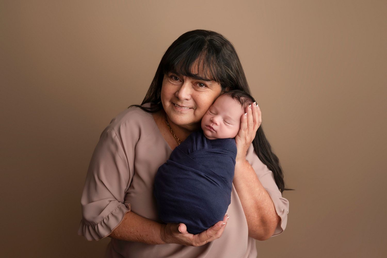 Woman with dark hair holds a sleeping newborn wrapped in navy blue swaddle against beige studio backdrop.