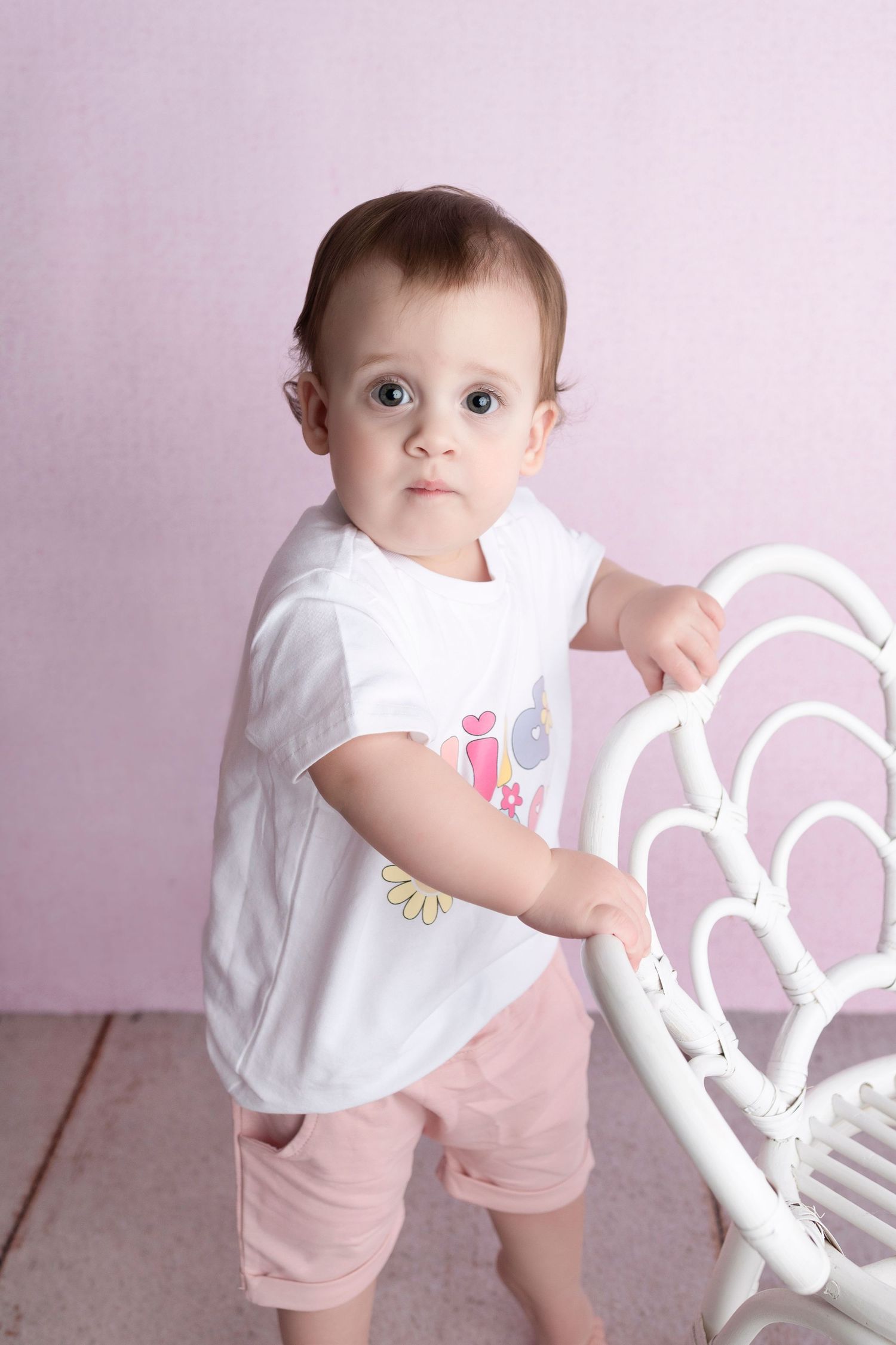 Cute baby in white graphic tee and pink shorts holding a white wicker chair on a light wood floor.