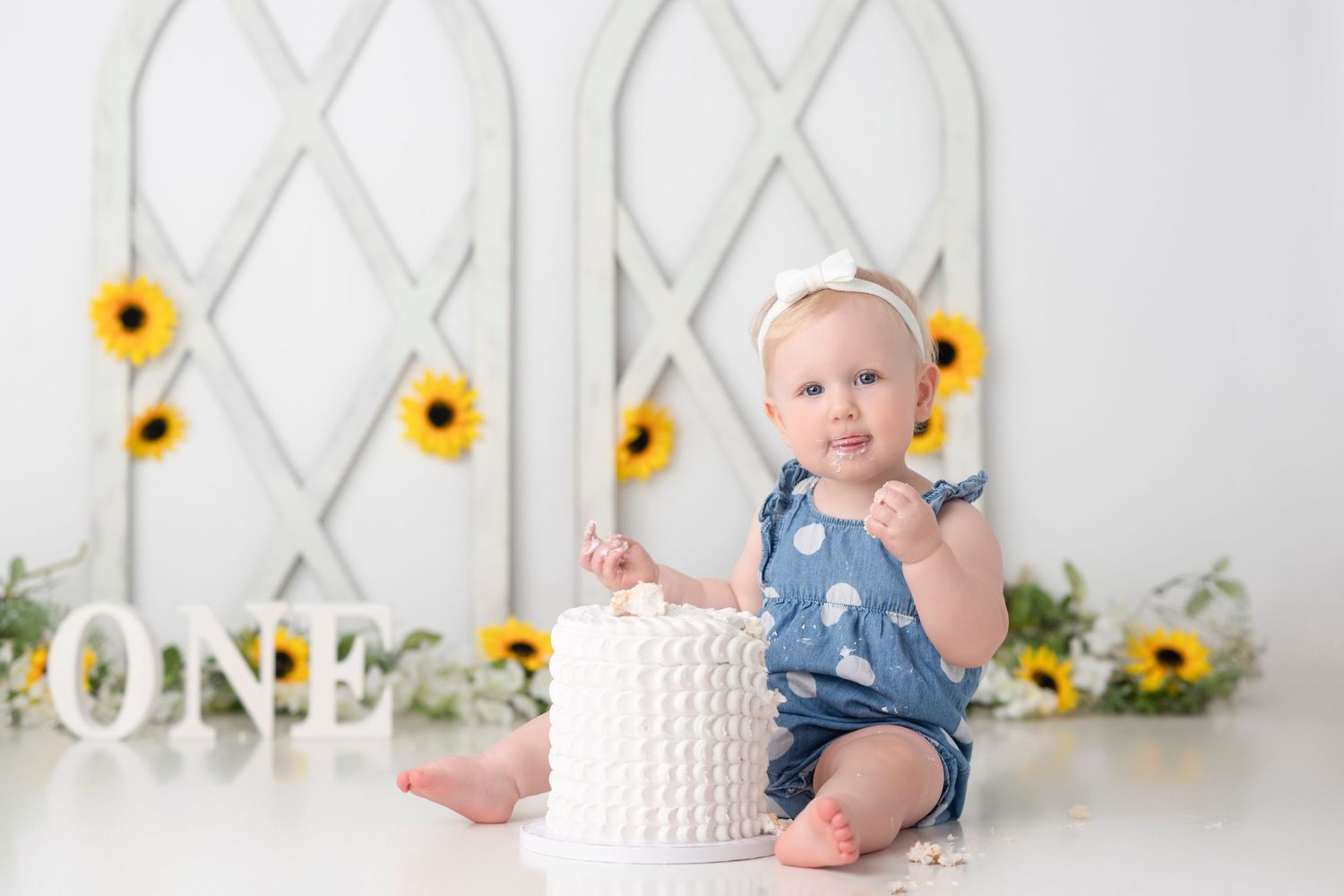 First birthday cake smash photo session with sunflowers and white lattice backdrop in a bright studio setting.