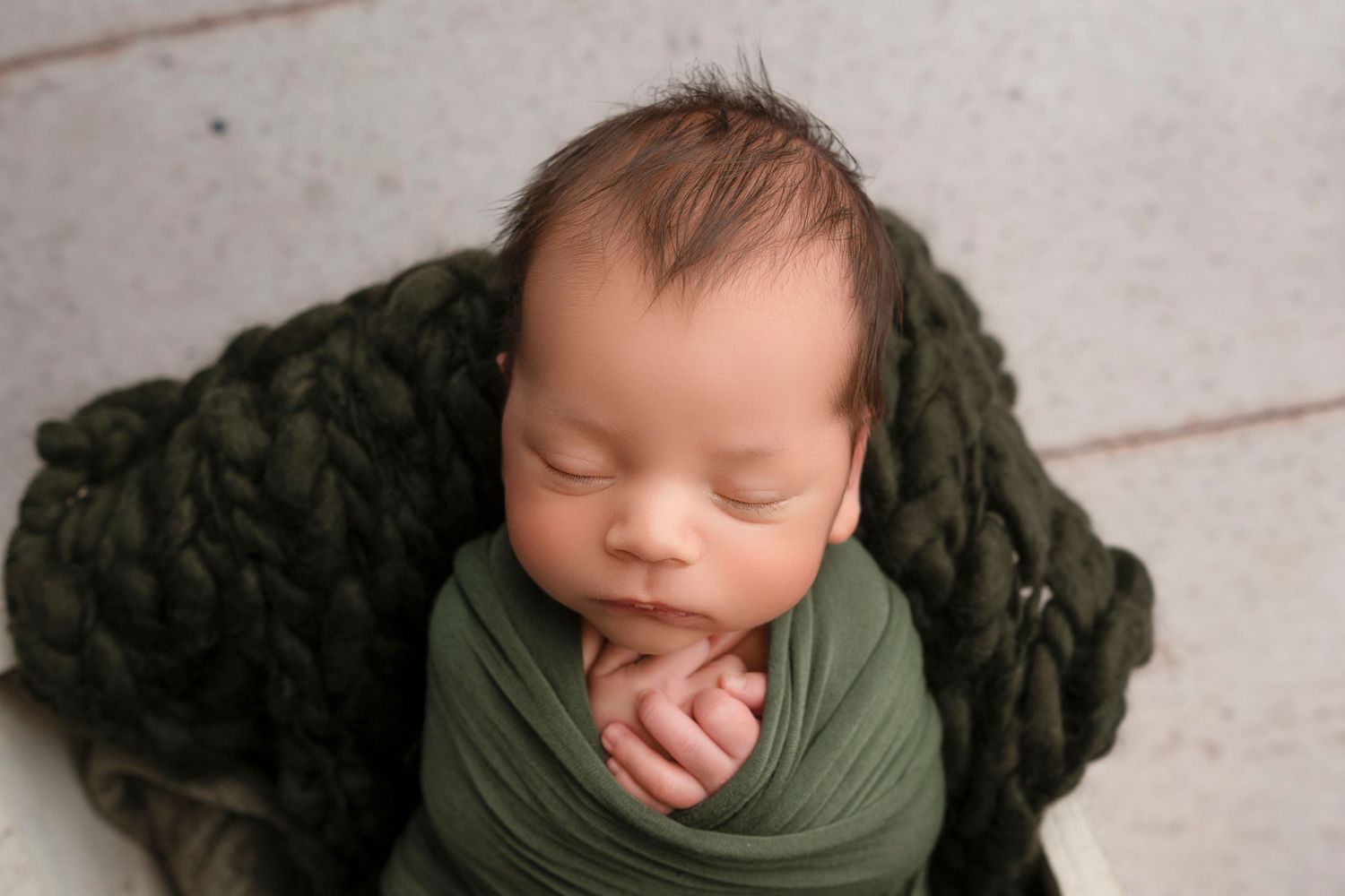 Newborn baby sleeping peacefully while wrapped in olive green swaddle blanket against wooden backdrop.