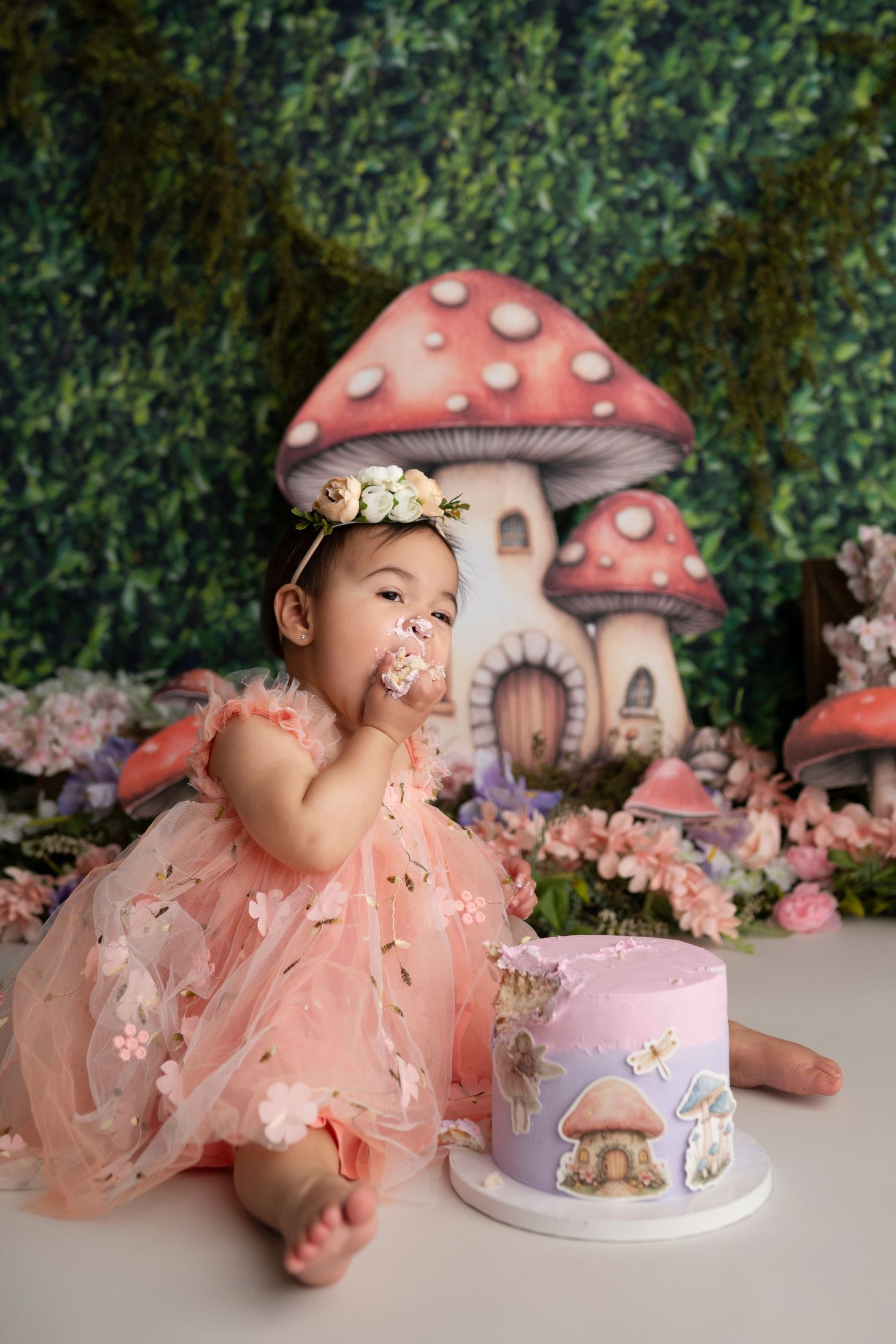 Whimsical woodland cake smash photo with pink dress and mushroom house backdrop featuring magical toadstool decorations.