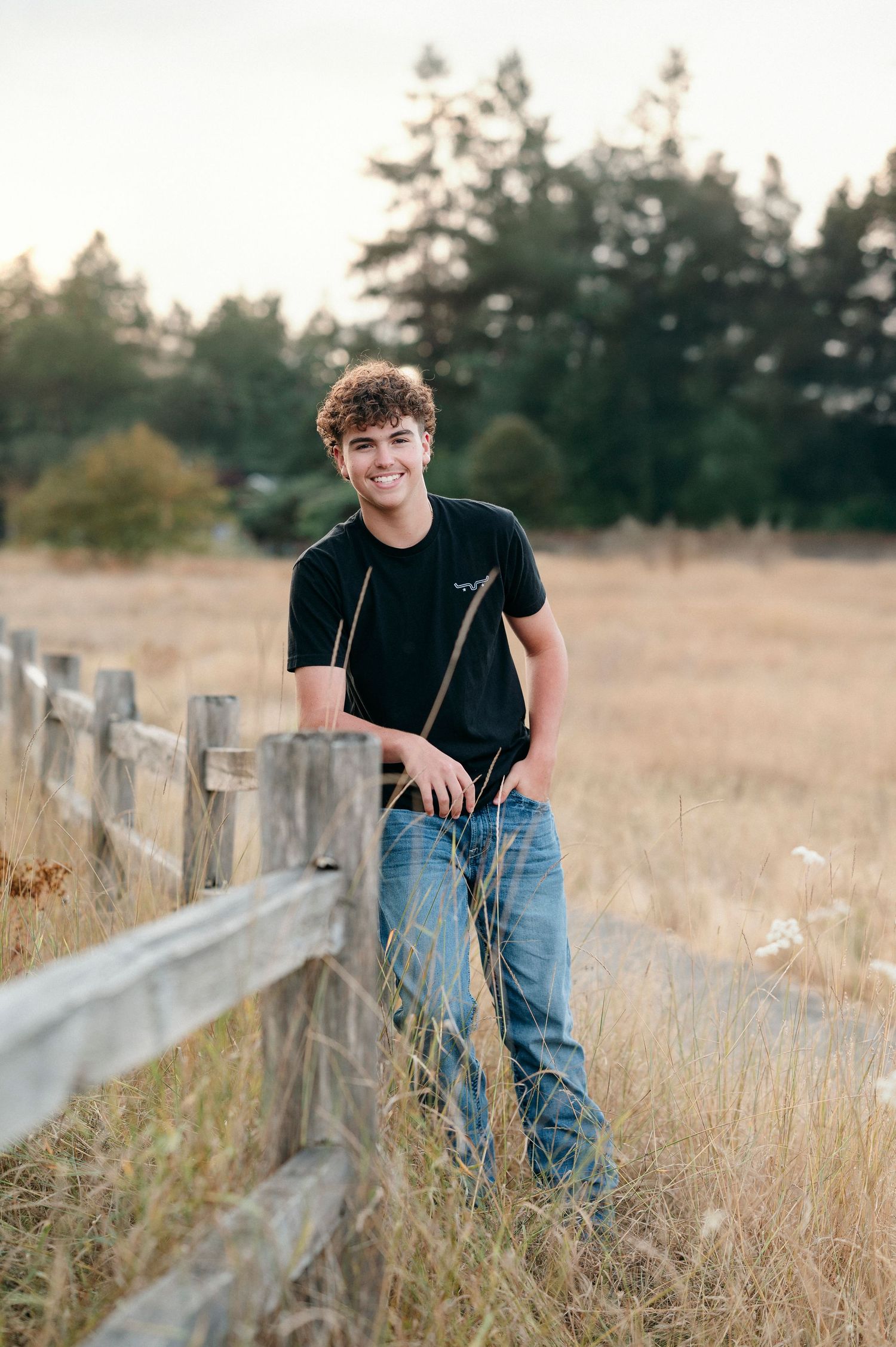 Casual outdoor portrait in a golden field with rustic wooden fence during sunset.