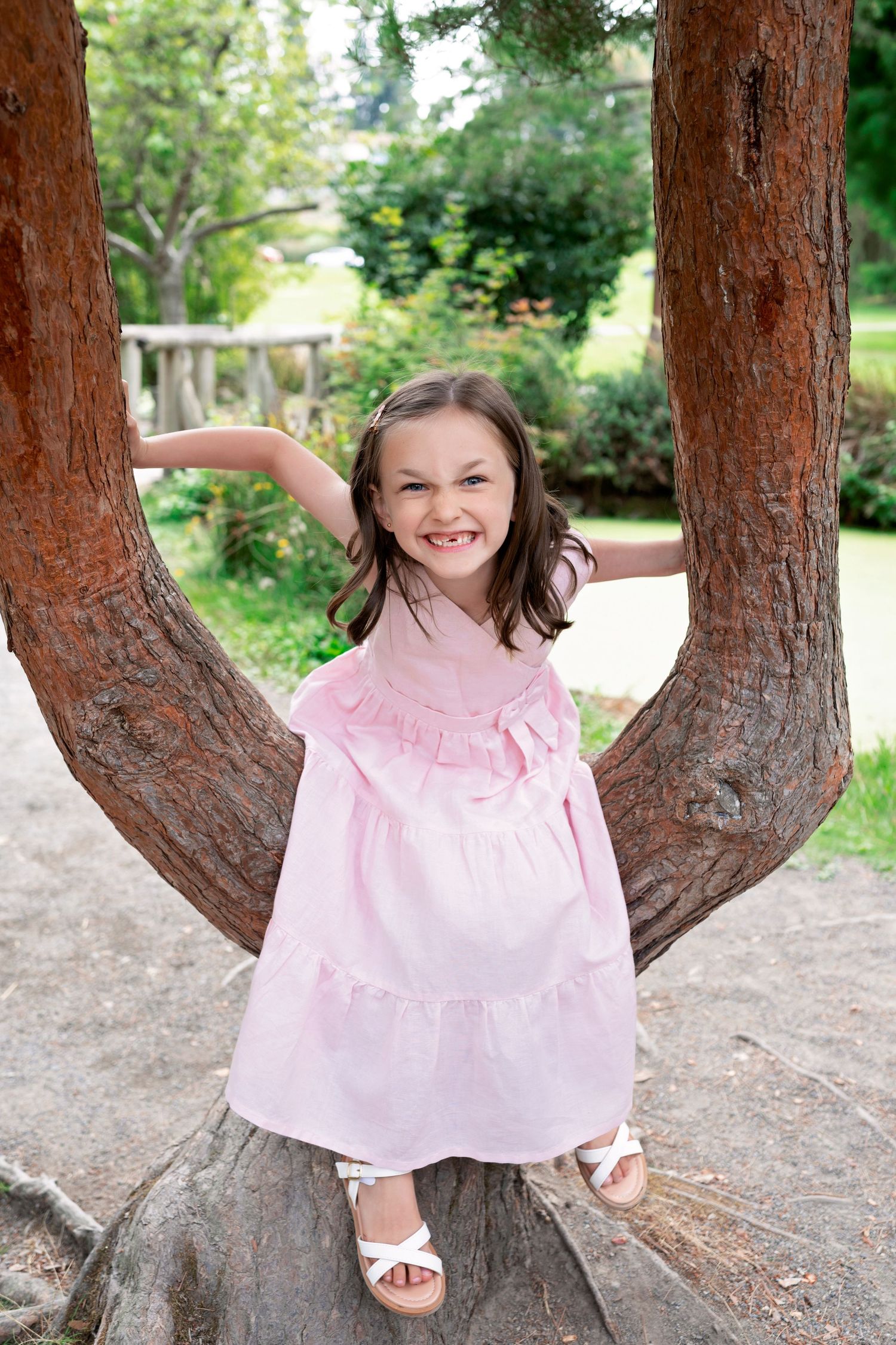 Child in pink dress plays on a branching tree trunk in a sunny garden setting.