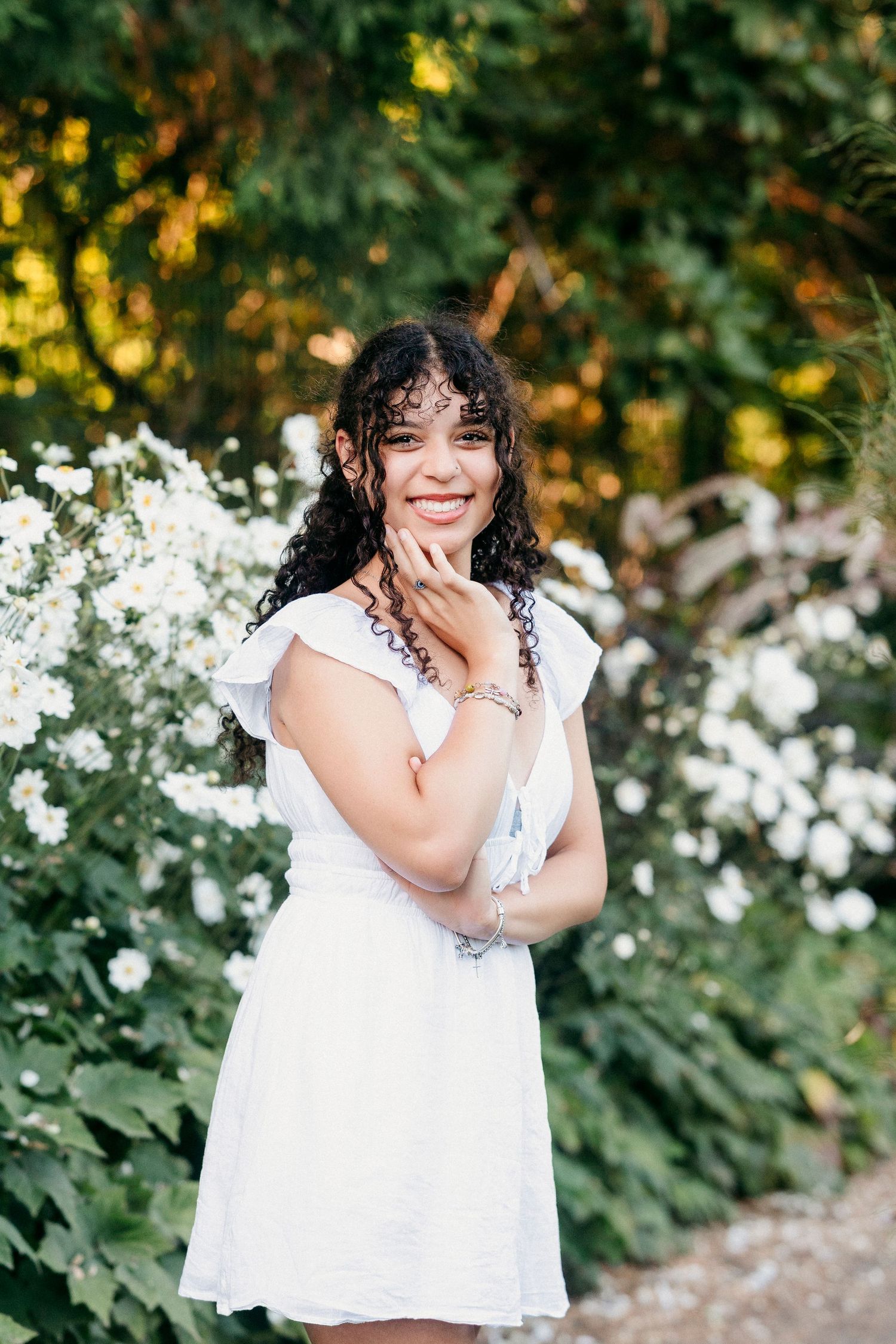 A person in a white summer dress stands smiling in front of white flowers and green foliage on a sunny day.