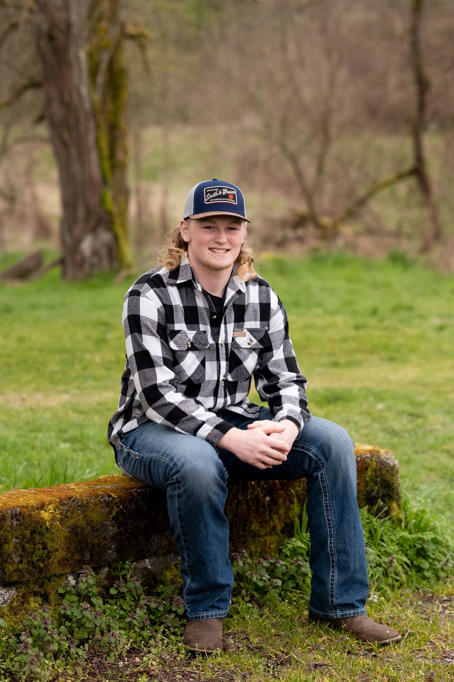 A person in a black and white plaid flannel shirt and jeans sits on a moss-covered log in a natural outdoor setting.