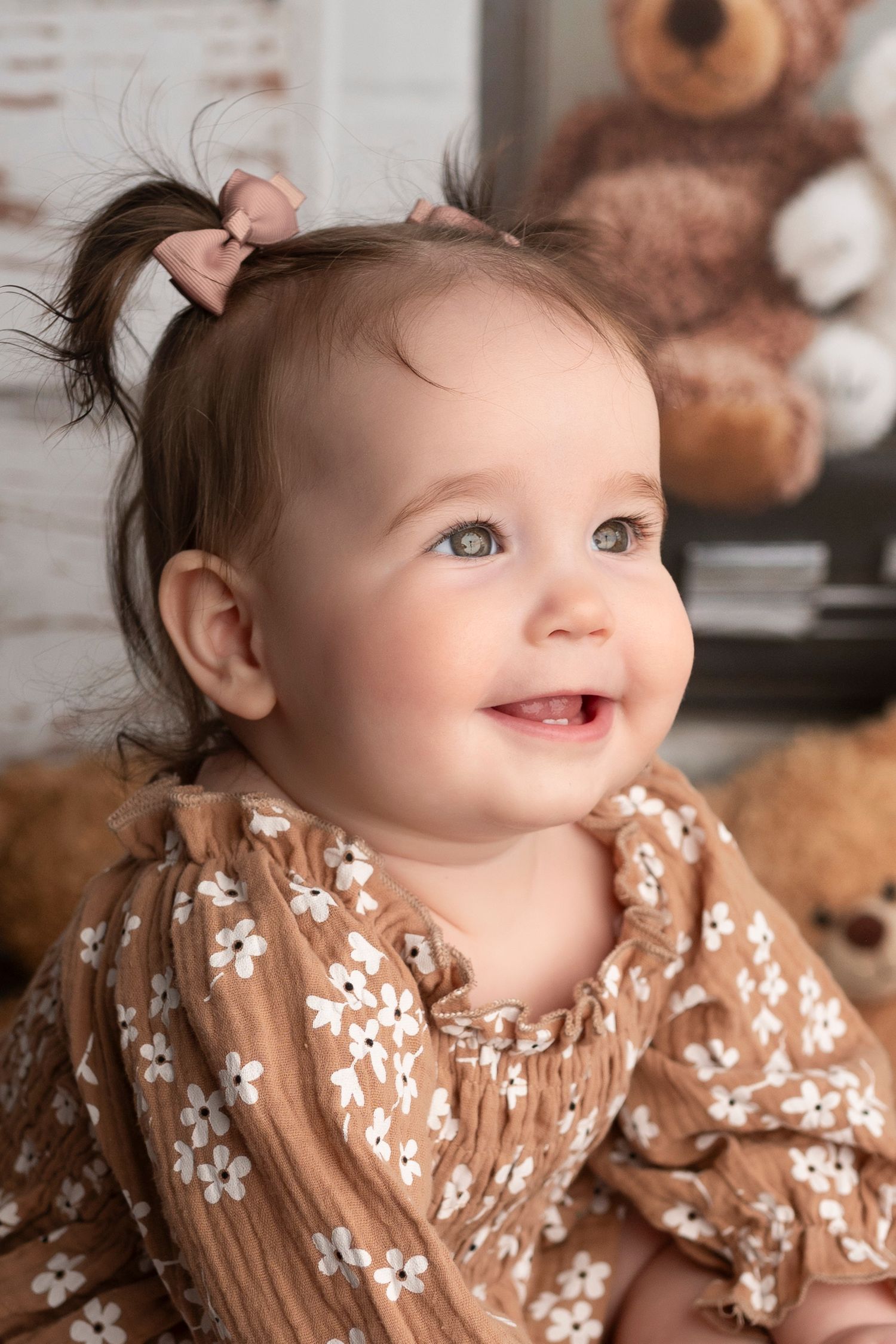 Smiling baby girl in a brown floral dress with a pink bow ponytail, sitting near a teddy bear.