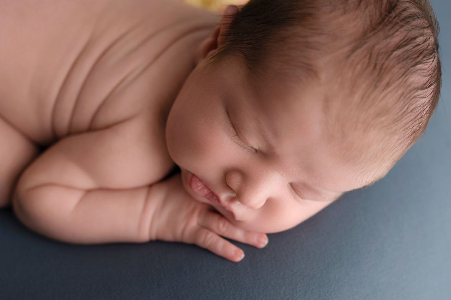Peaceful newborn baby sleeping on a blue surface, with tiny hands tucked under cheek in a professional portrait photo.