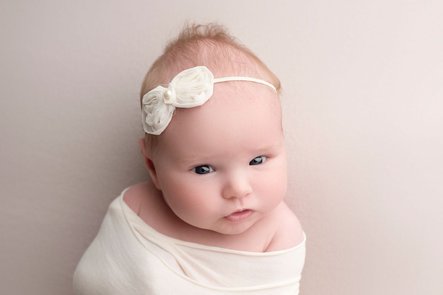Newborn baby girl wrapped in white swaddle cloth wearing a delicate white bow headband on neutral background.