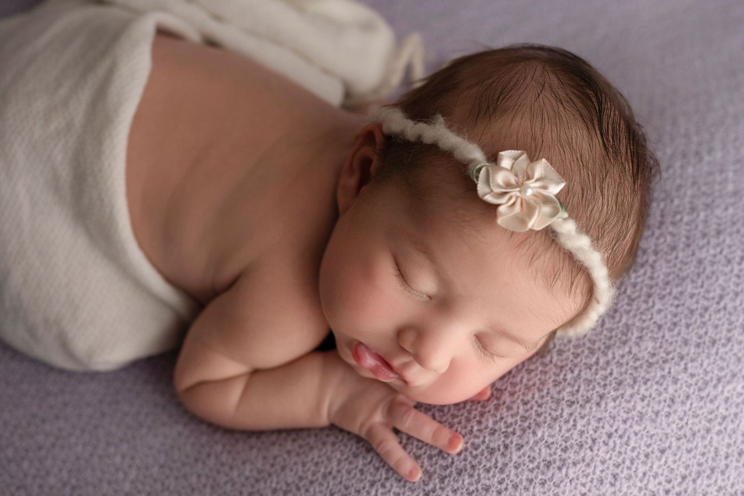 Peaceful newborn baby girl sleeping wrapped in white, wearing a delicate floral headband on a soft lavender blanket.