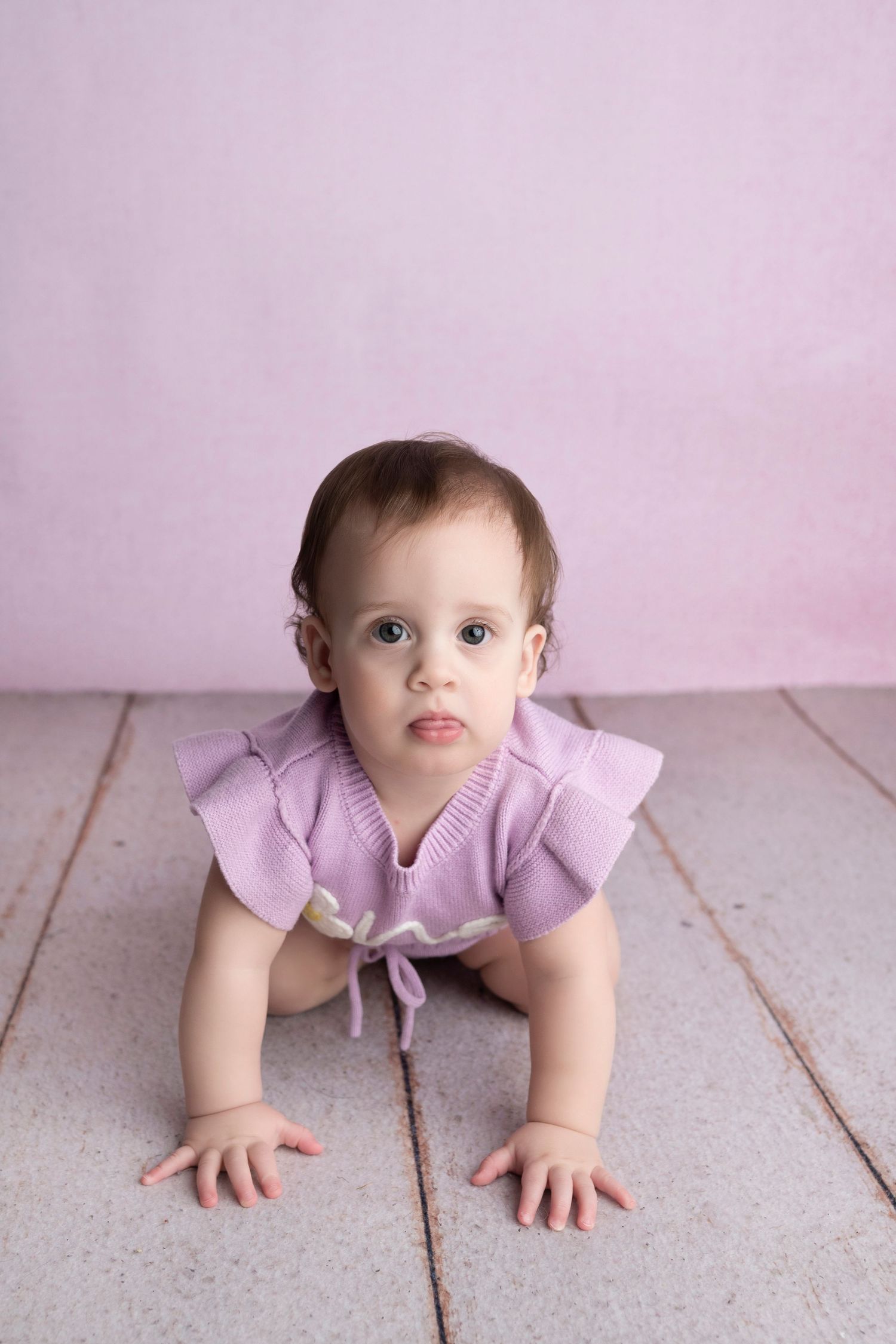 A baby girl in a purple ruffle romper crawls on a wooden floor against a soft pink background.