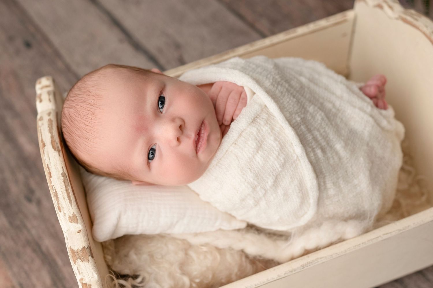 Newborn baby swaddled in cream wrap lying in a rustic white wooden crate with soft fur lining.