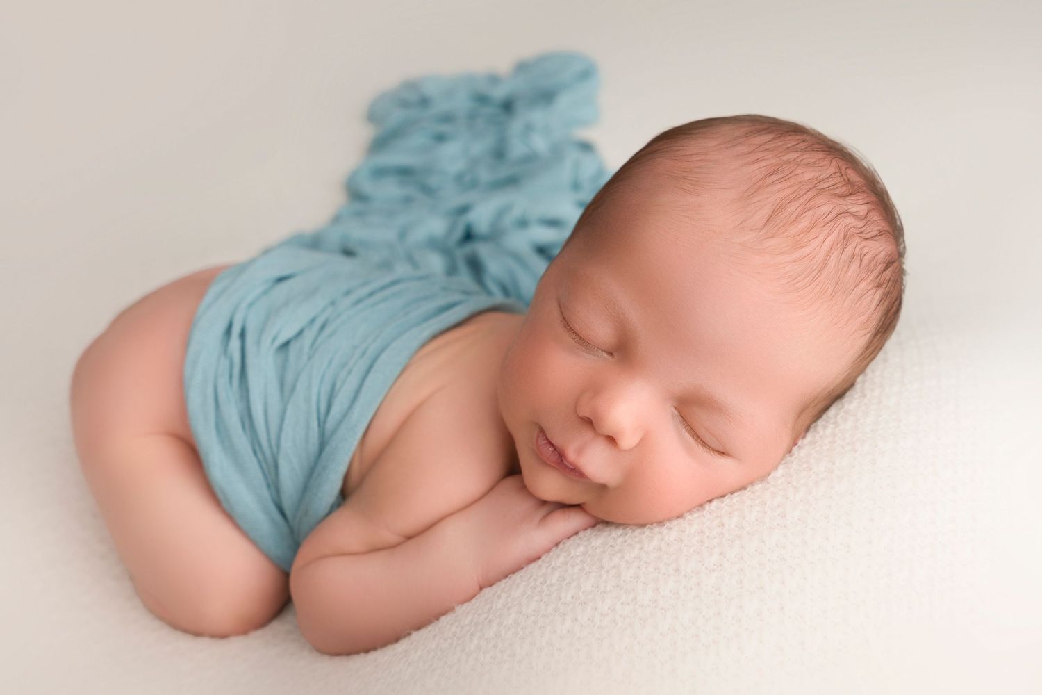 Sleeping newborn baby wrapped in soft turquoise fabric lying on white backdrop during professional photo session.