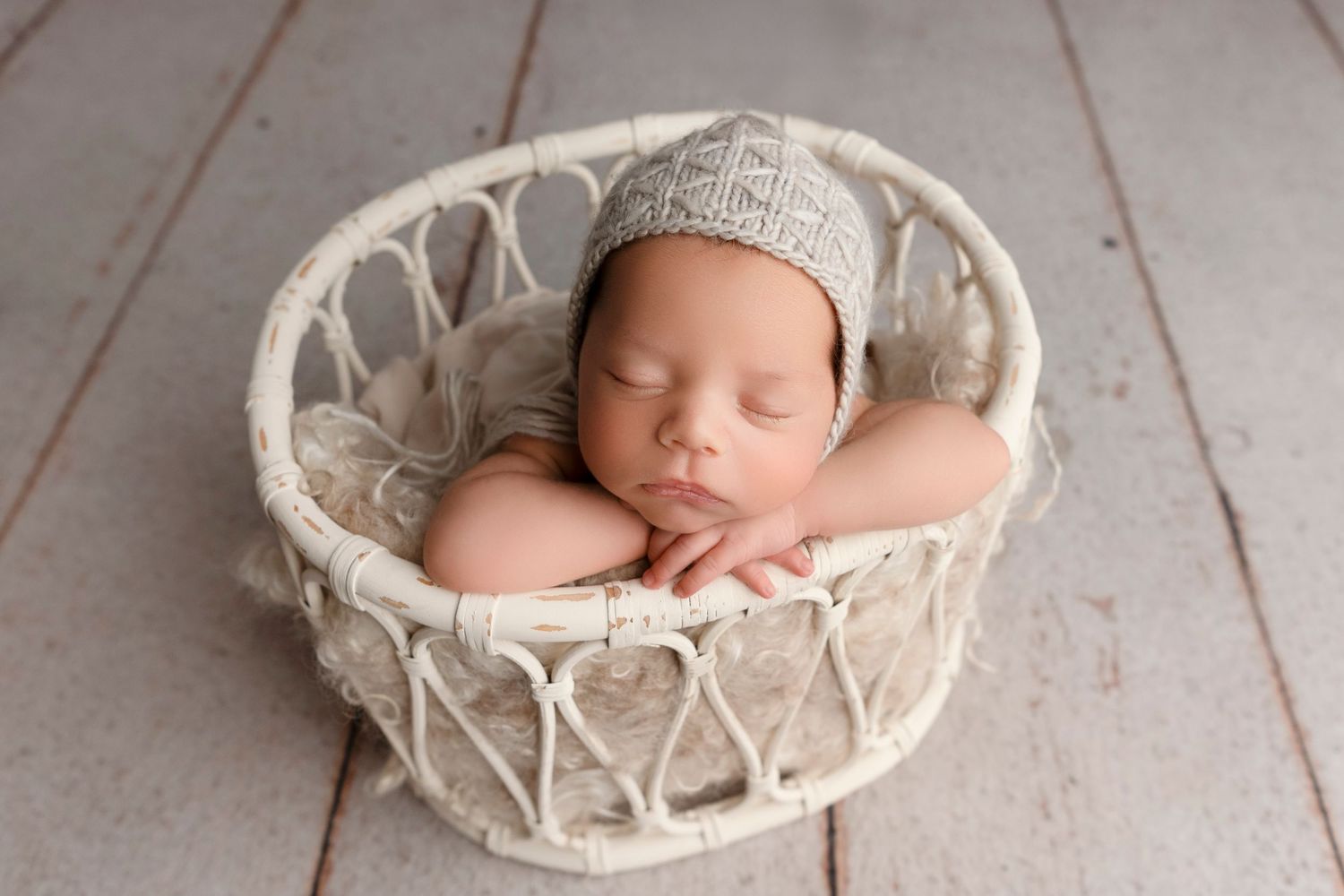 Sleeping newborn baby wrapped in cream-colored blanket rests peacefully in woven basket on wooden floor.