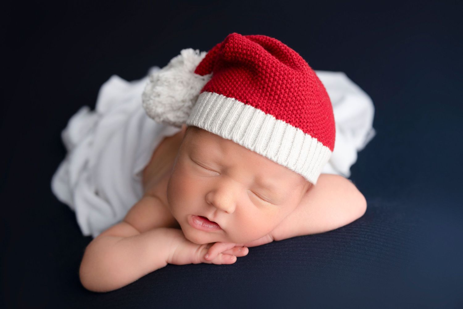 A sleeping newborn wearing a red and white knit hat against a dark blue backdrop.