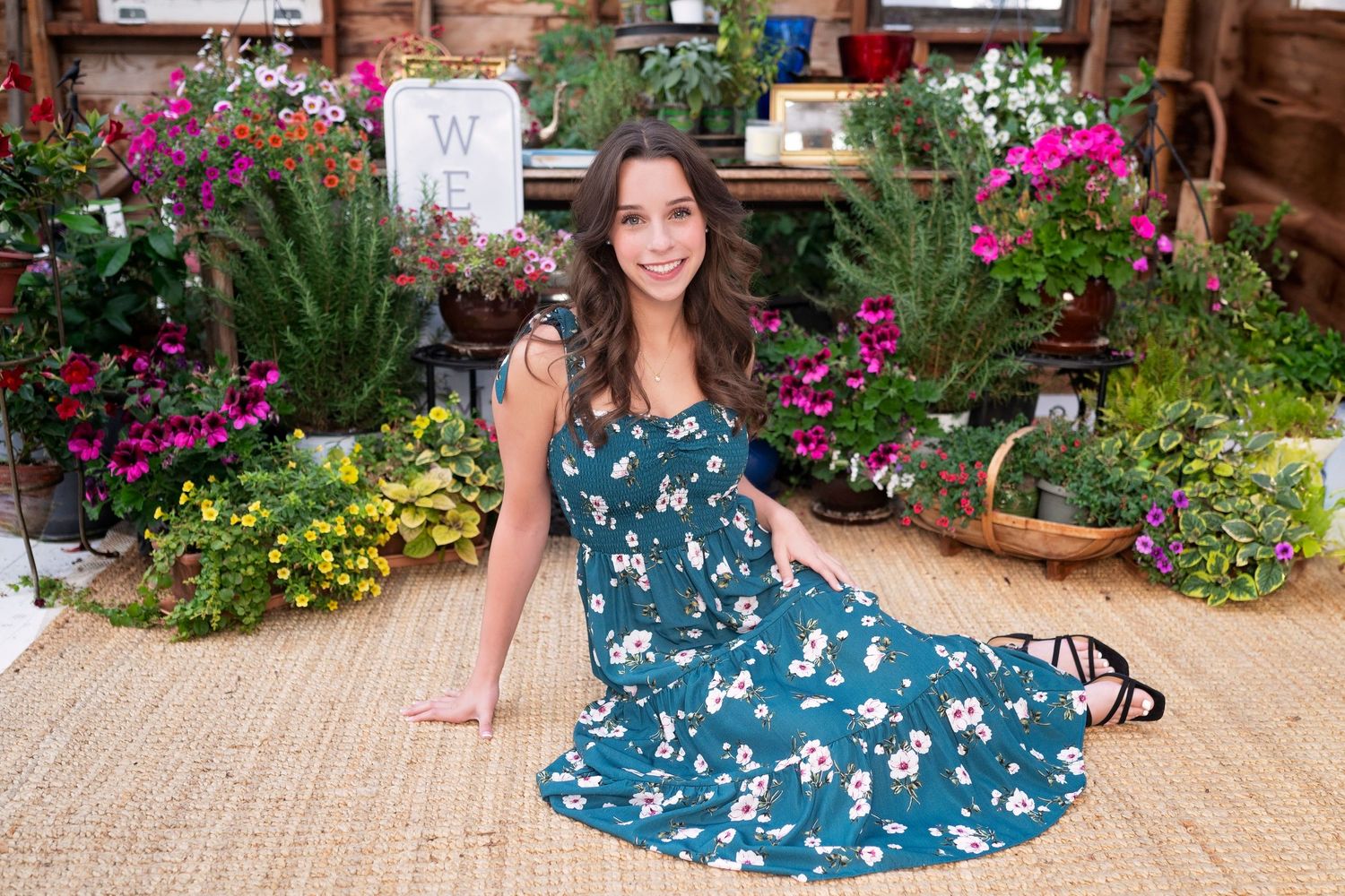 Woman in floral teal dress sitting on woven mat surrounded by colorful garden plants and flowers.