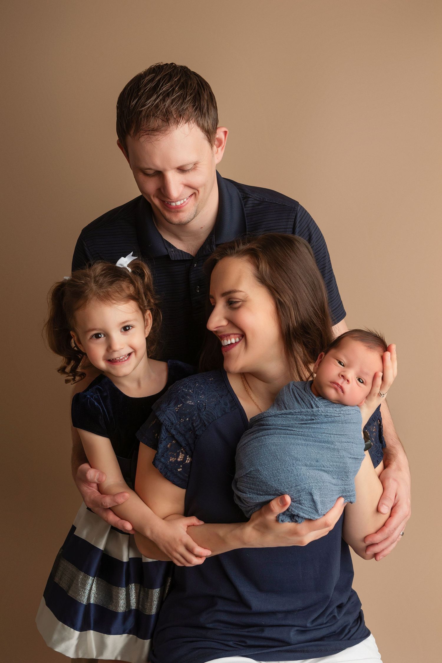 A smiling family of four poses together, parents holding a toddler girl and newborn baby wrapped in blue.